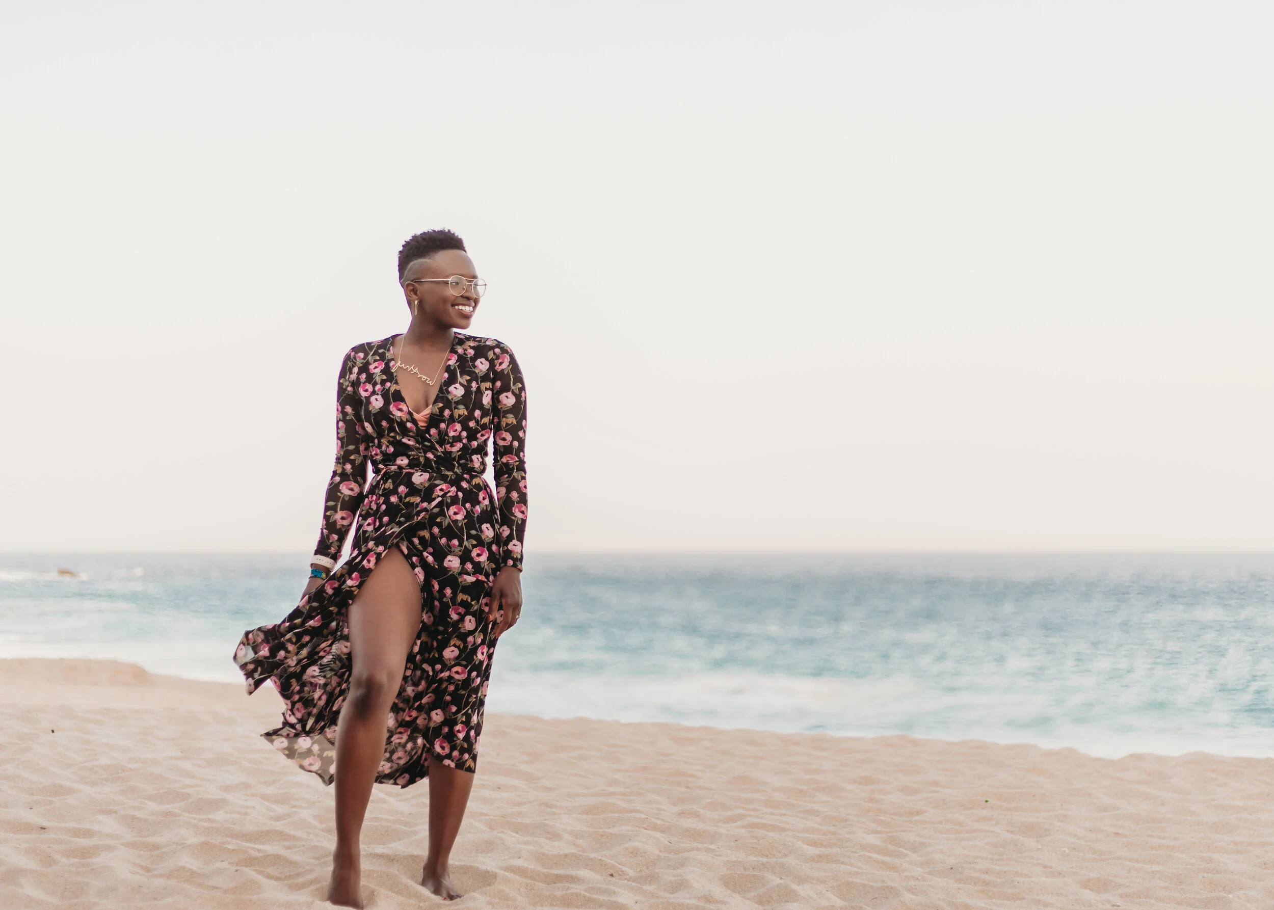 A woman in a floral dress walking on the beach, smiling, with the ocean in the background.