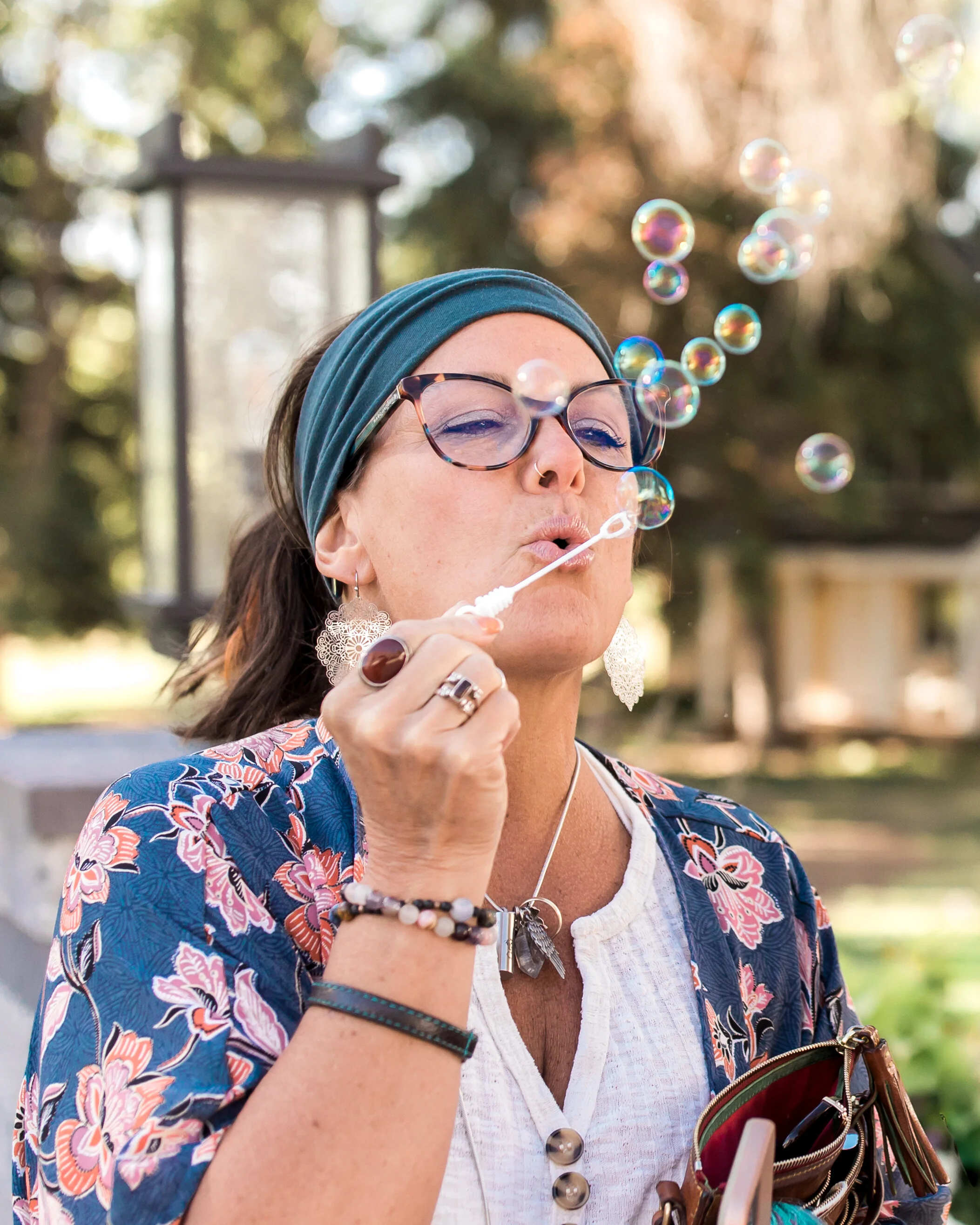 A woman blowing bubbles outdoors on a sunny day, wearing glasses, a headscarf, and colorful clothing with various accessories.