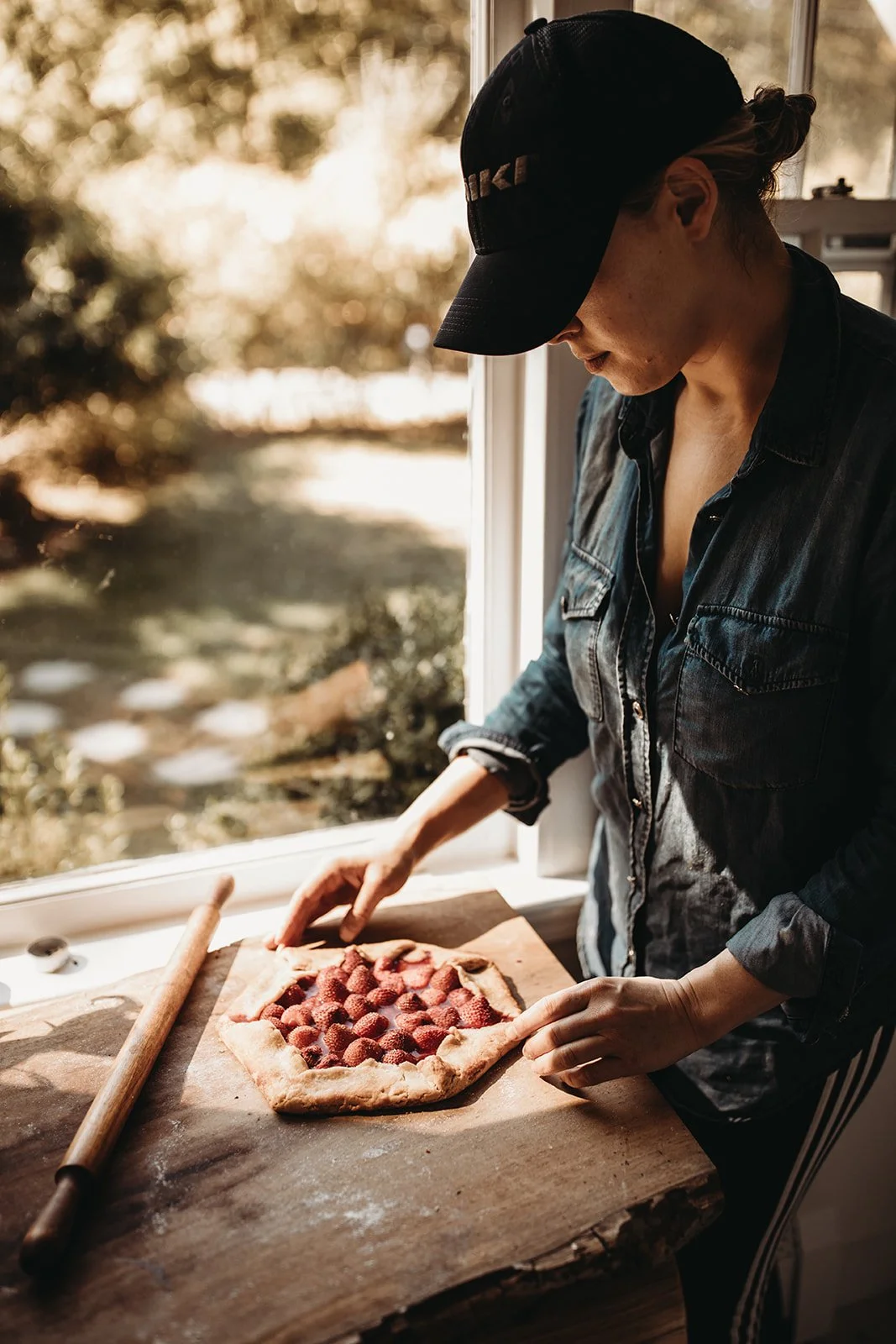A woman in a black denim jacket and cap making a strawberry galette in a sunlit kitchen.