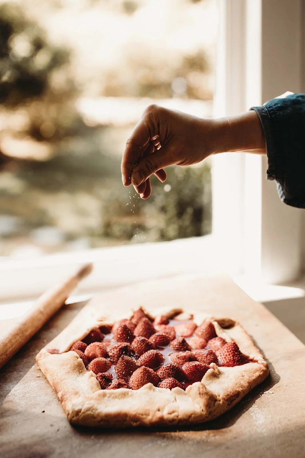 Woman sprinkling sugar over a freshly assembled strawberry galette on a baking sheet near a window.