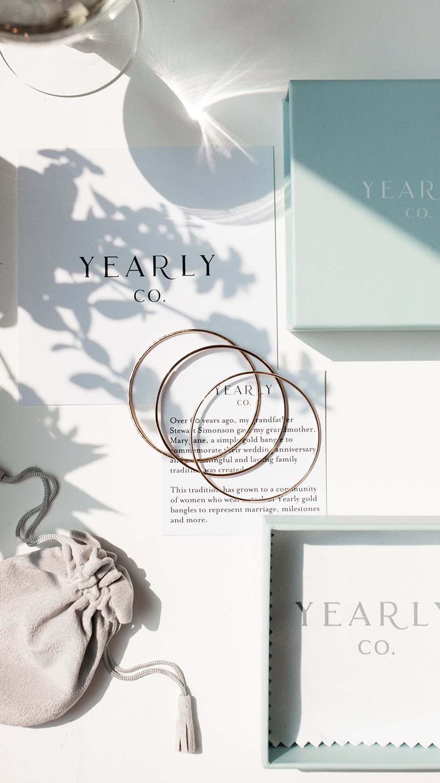 Three gold bangles, a small beige velvet pouch, a jewelry box, and informational cards from Yearly Co. on a white surface with natural sunlight and shadows.
