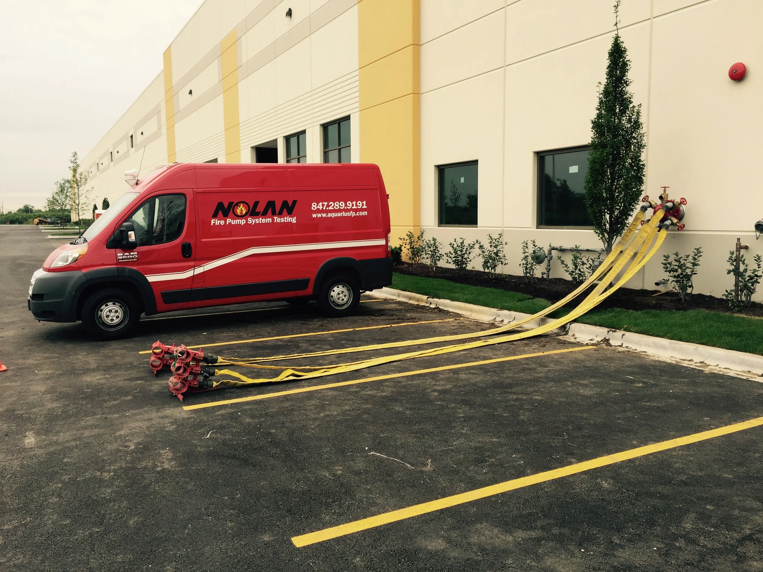 Red van labeled 'NOLAN Fire Pump System Testing' parked in an empty parking lot next to a building that has a test header. Fire hose and hose monsters are connected and set up ready to perform a fire pump flow test.