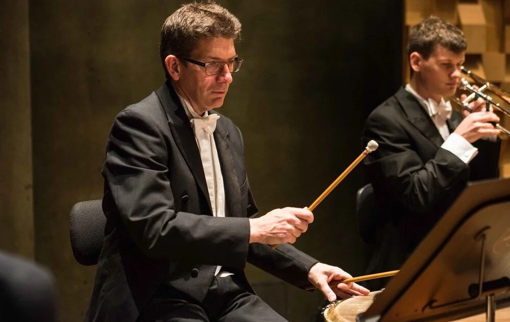 Two musicians in formal attire performing in an indoor setting. The man in the foreground is playing percussion with drumsticks, and the man behind him is playing the trumpet.