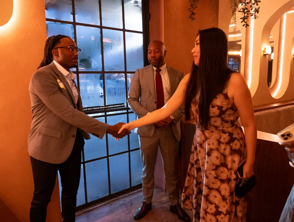 A woman in a floral dress shaking hands with a man in a gray suit inside a dimly lit restaurant or bar, with two other men in suits observing.