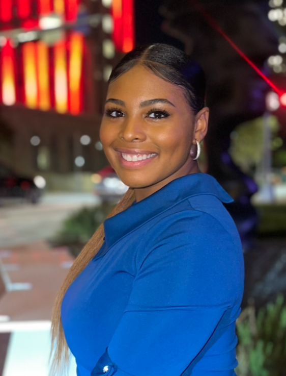 A smiling woman with dark hair styled in a sleek ponytail, wearing hoop earrings and a blue top, standing outdoors at night with blurred colorful lights in the background.