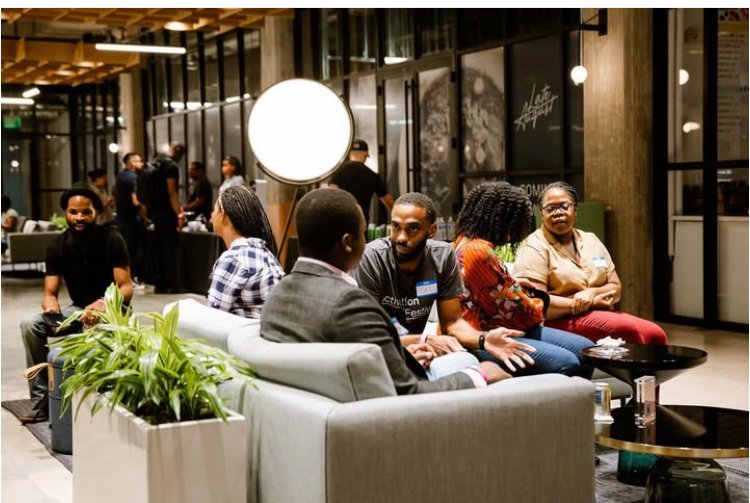 A group of people sitting and talking in a modern lounge or waiting area with glass walls, potted plants, and contemporary decor.