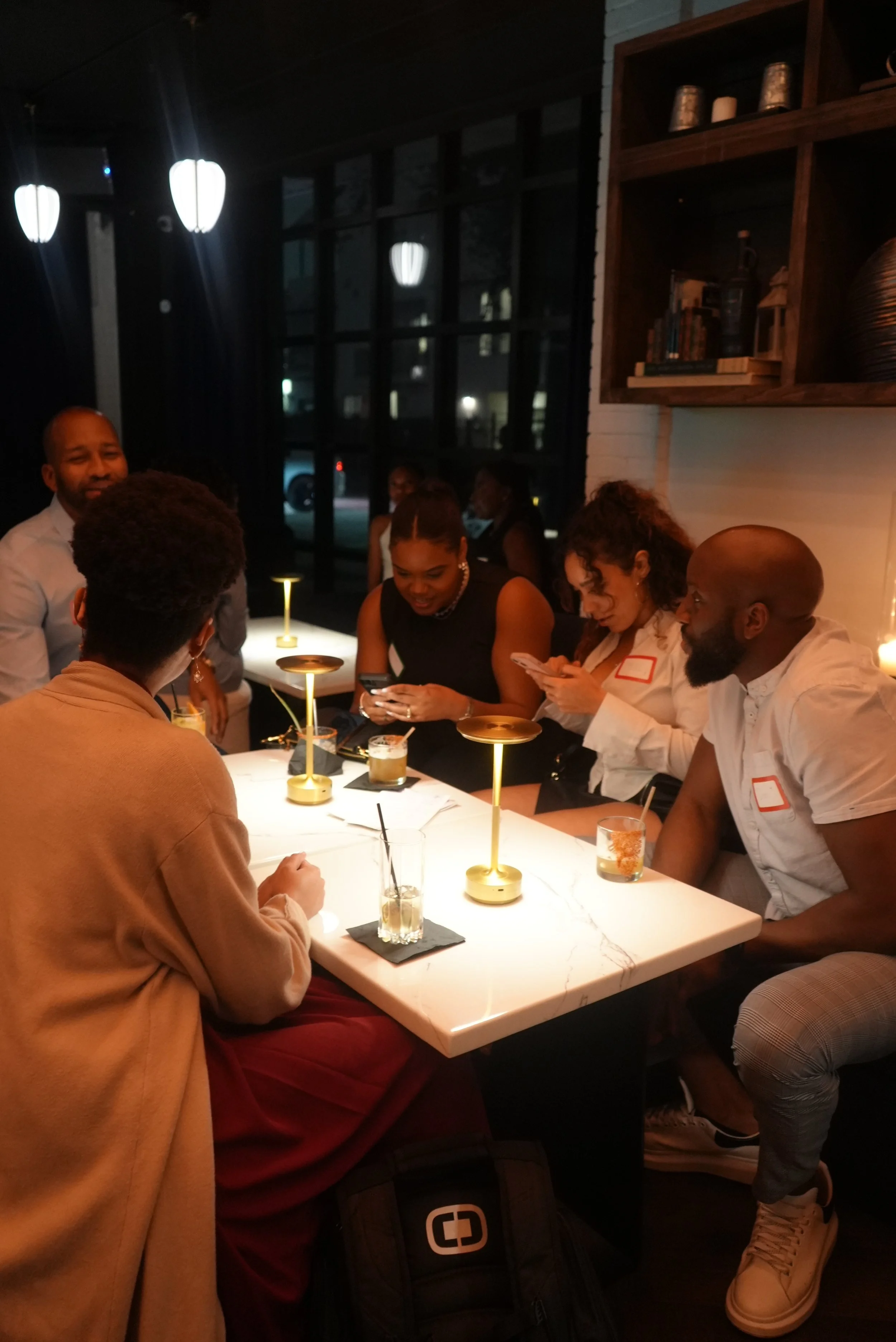 A group of seven people gathered around a white marble table in a dimly lit restaurant, engaging in conversations and looking at their phones, with modern lamps and decor in the background.