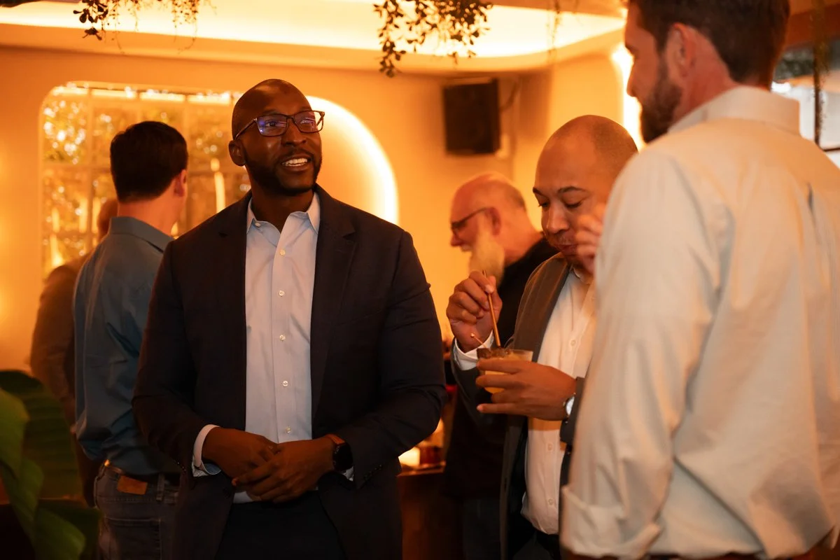 People socializing at a party or event in a warm, cozy setting, with three men engaged in conversation, one of whom is drinking from a glass with a straw.