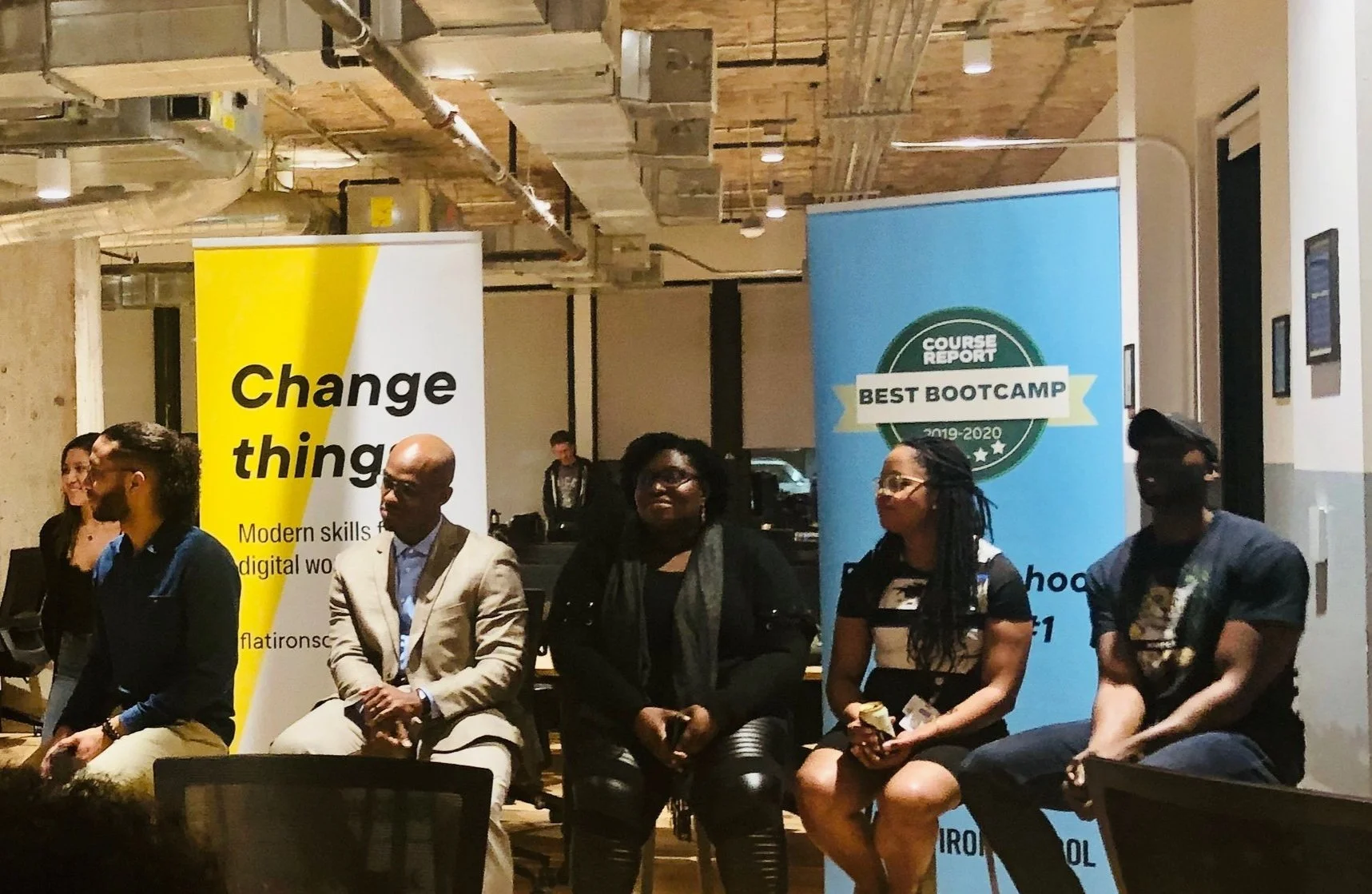 Group of six diverse people sitting on chairs at a panel discussion or event in a modern, industrial-style room with banners promoting skill development and a bootcamp.