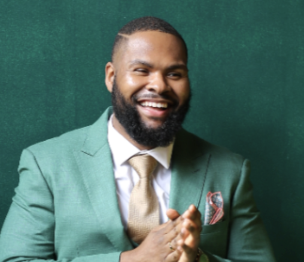 Portrait of a smiling African American man dressed in a green suit, white shirt, and gold tie, posed against a green background.