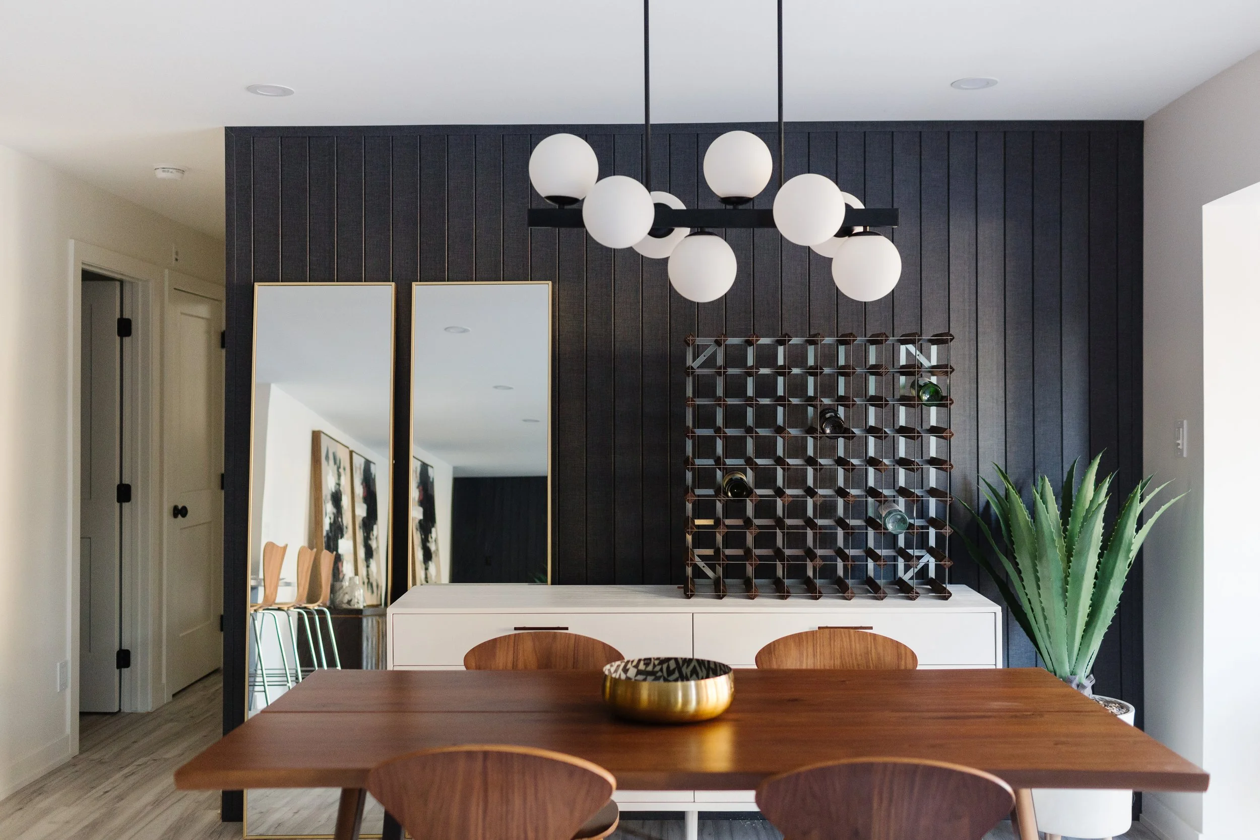 Photo of a black accent wall in a modern dining room with mirrors, a wine rack, a white side table, and a houseplant behind a wood dining table and chairs
