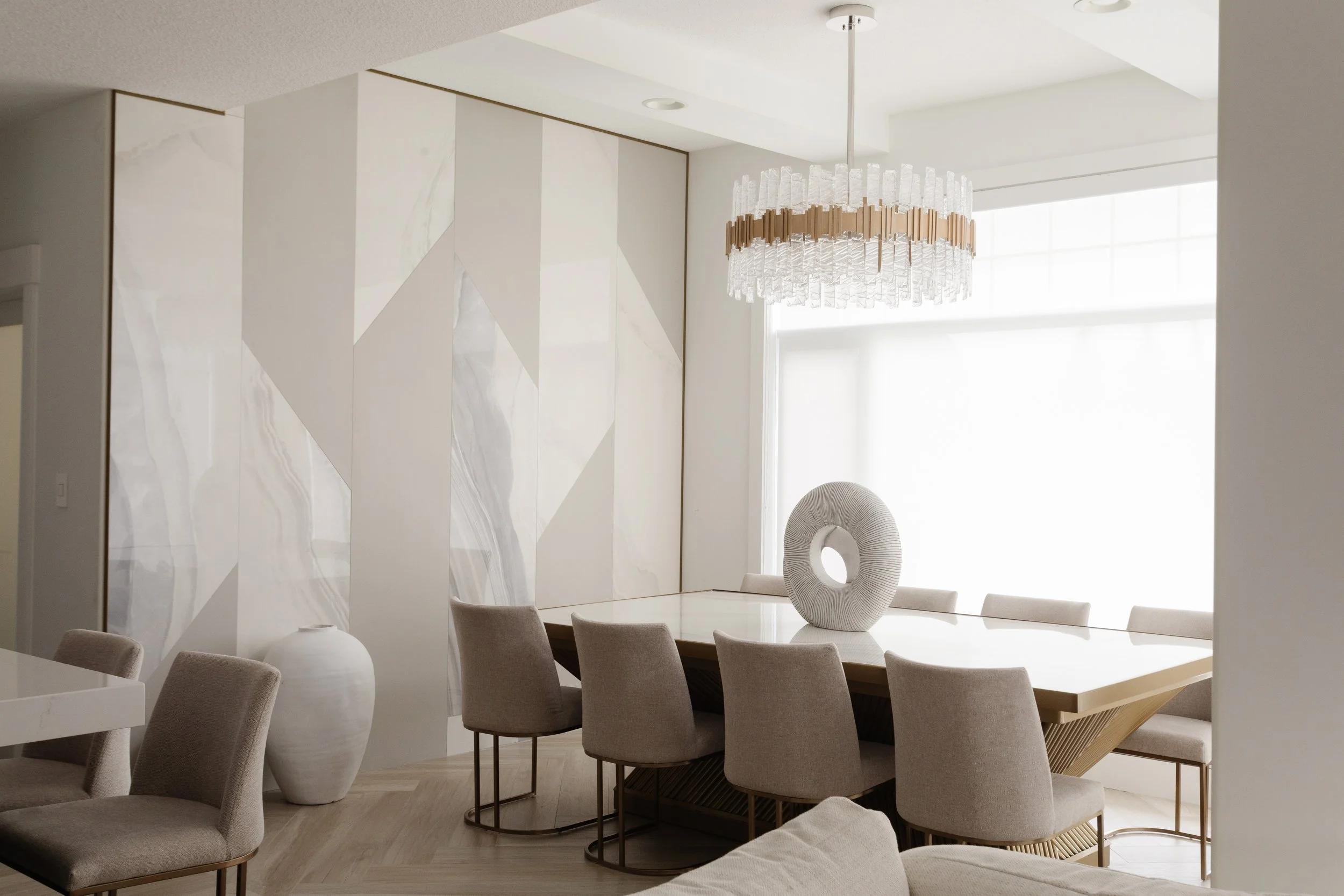 Photo of a white luxury dining room with a glass chandelier over top of a gold dining table with beige dining chairs and white home decor