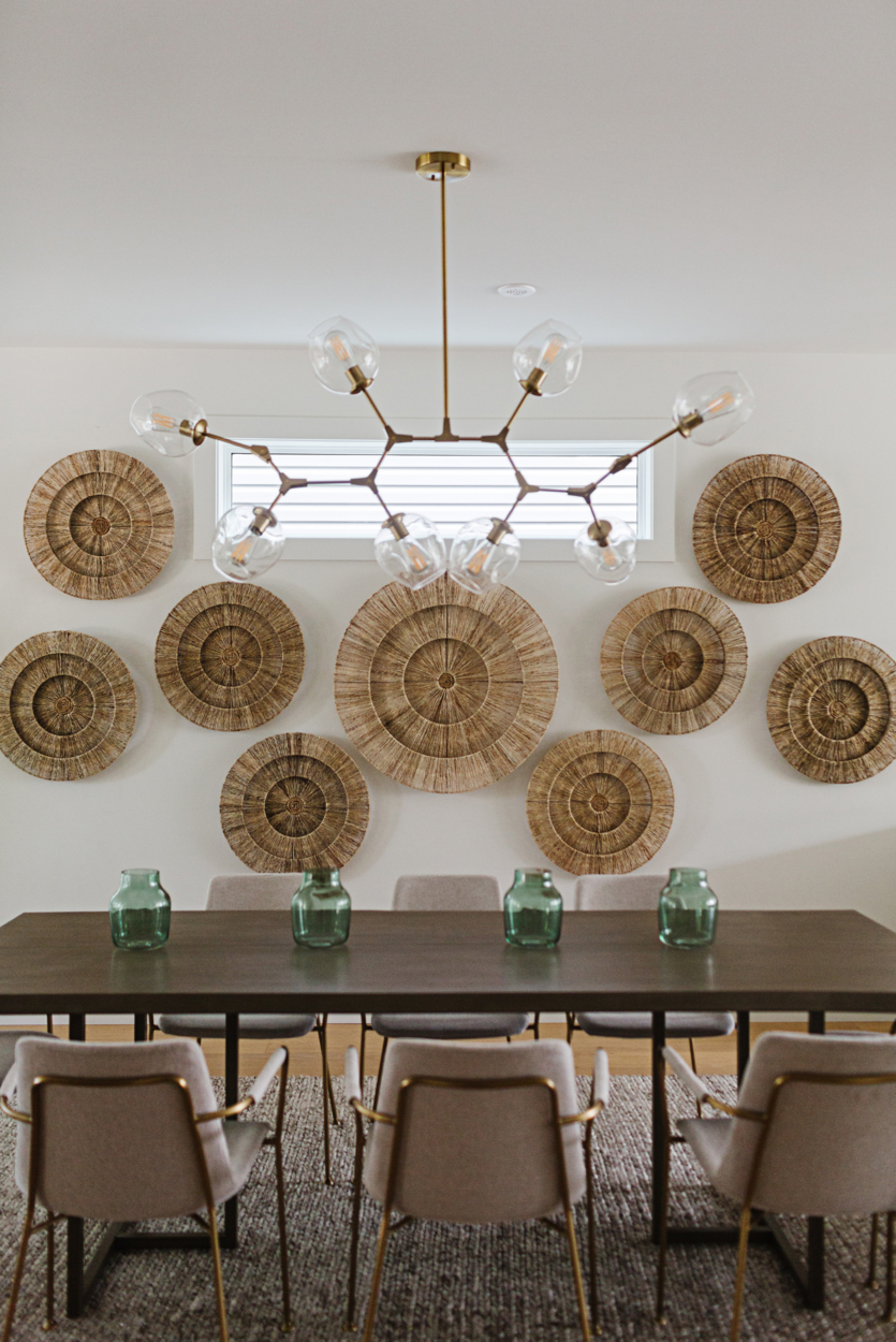 Photo of a dining room table and chairs beneath a gold light bulb fixture with large, round wood decor hanging on the wall