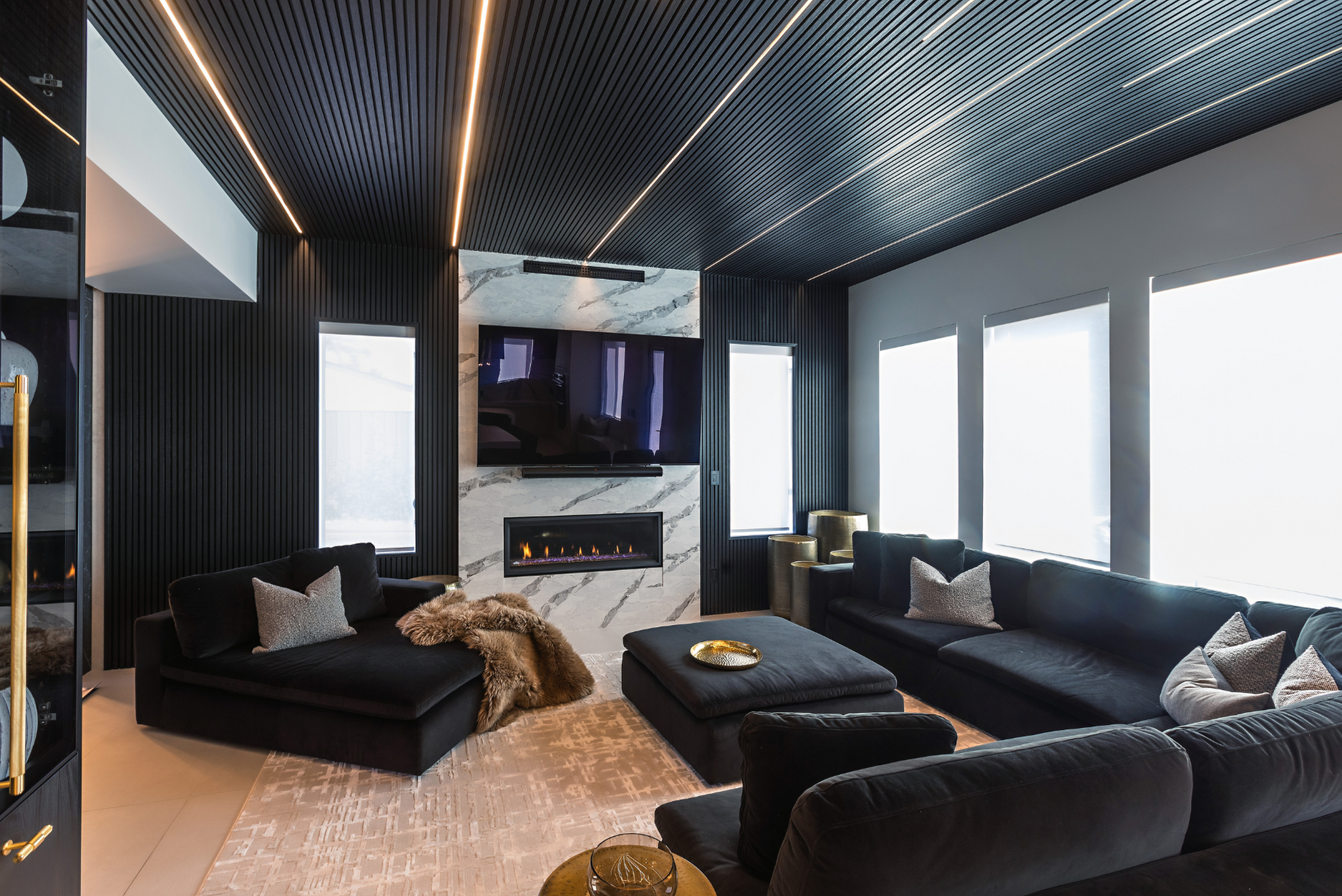 Picture of a modern living room, demonstrating how to design a multifunctional living space, featuring a black slatted ceiling with integrated linear lighting, a white marble fireplace wall with a built-in TV, and a large black sectional sofa