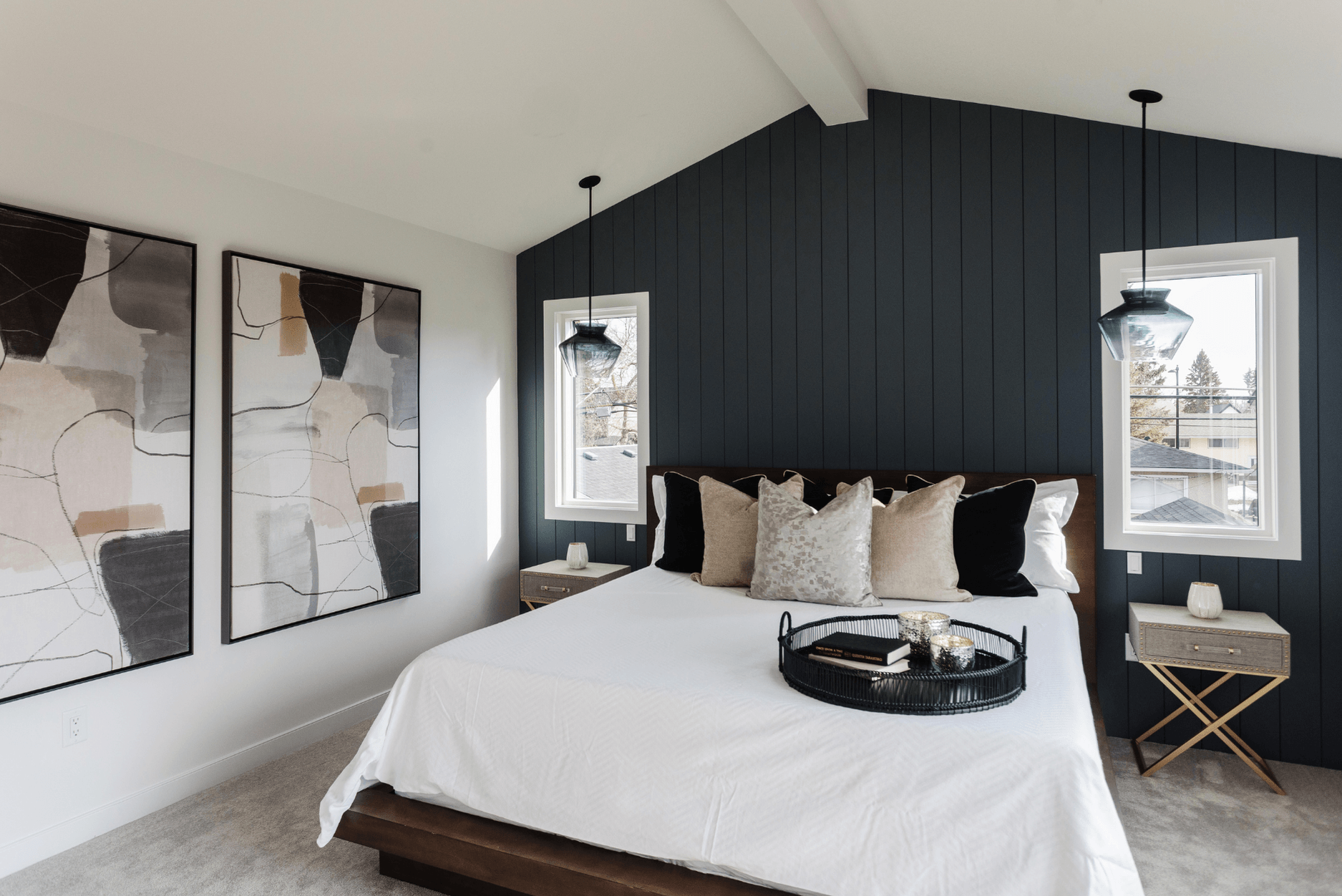 A photo of a stylish bedroom featuring a dark vertical panelled accent wall, abstract artwork, pendant lights, and a white bed with layered neutral pillows