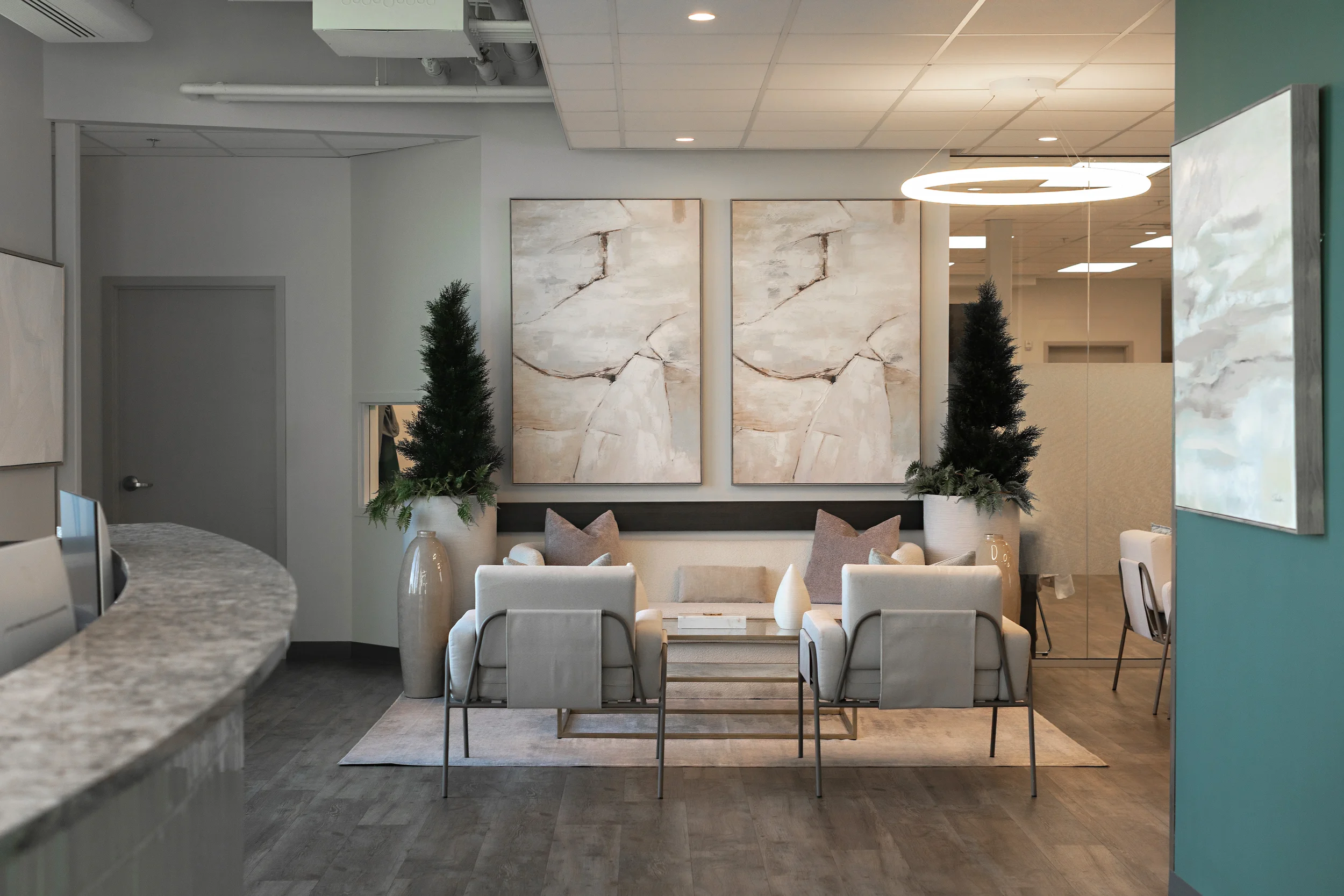 photo of two white armchairs in a waiting room facing a white sofa below two matching artworks and two pine trees
