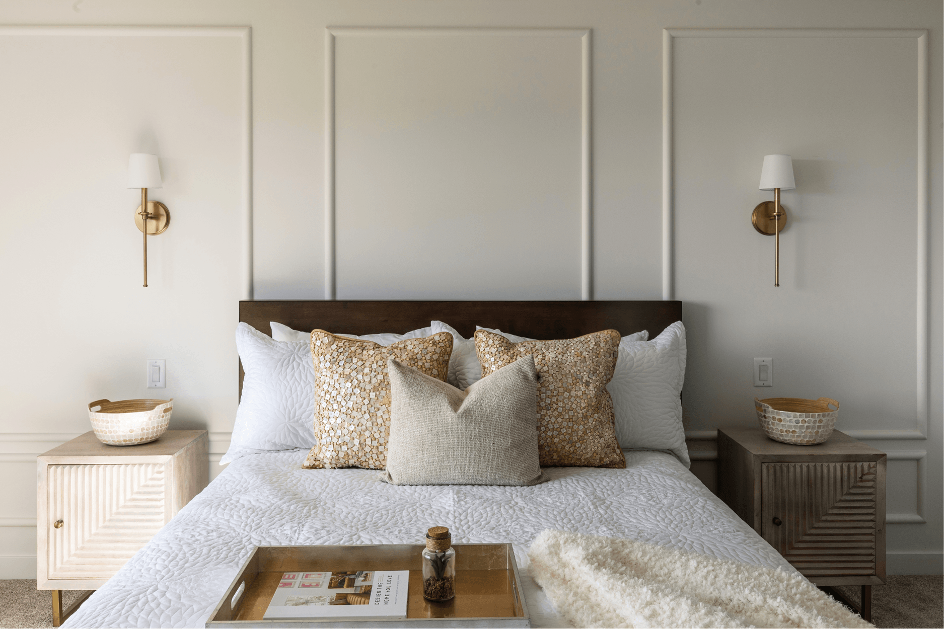 Photo of a bedroom with a white bed, brown throw pillows, a tray, and a blanket next to matching wood cube nightstands below gold wall lamps