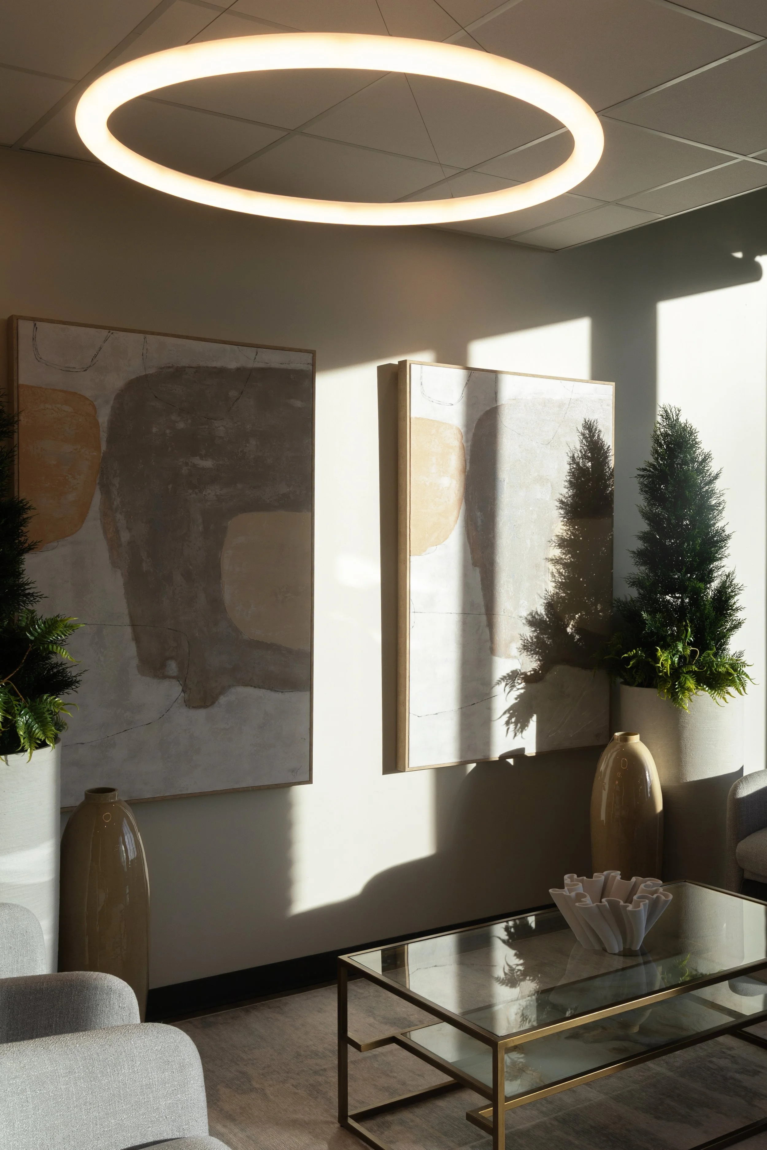 Photo of a room with matching abstract artwork next to pine tree and decor on a glass coffee table below a circular light fixture
