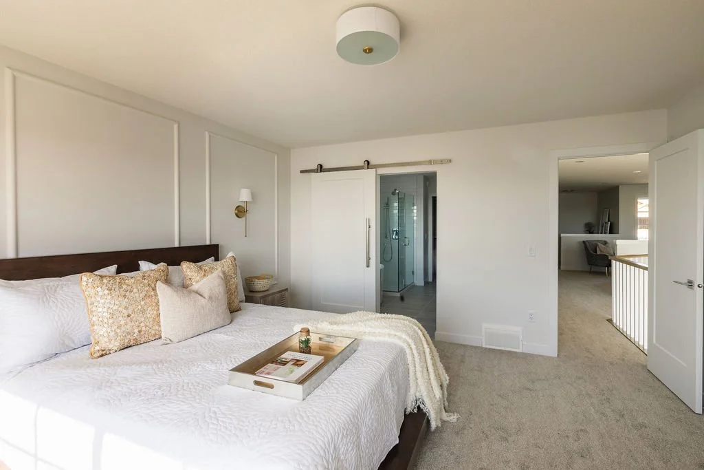 A photo of a cozy neutral bedroom with white bedding soft carpeting and a sliding barn door opening to a glass shower ensuite
