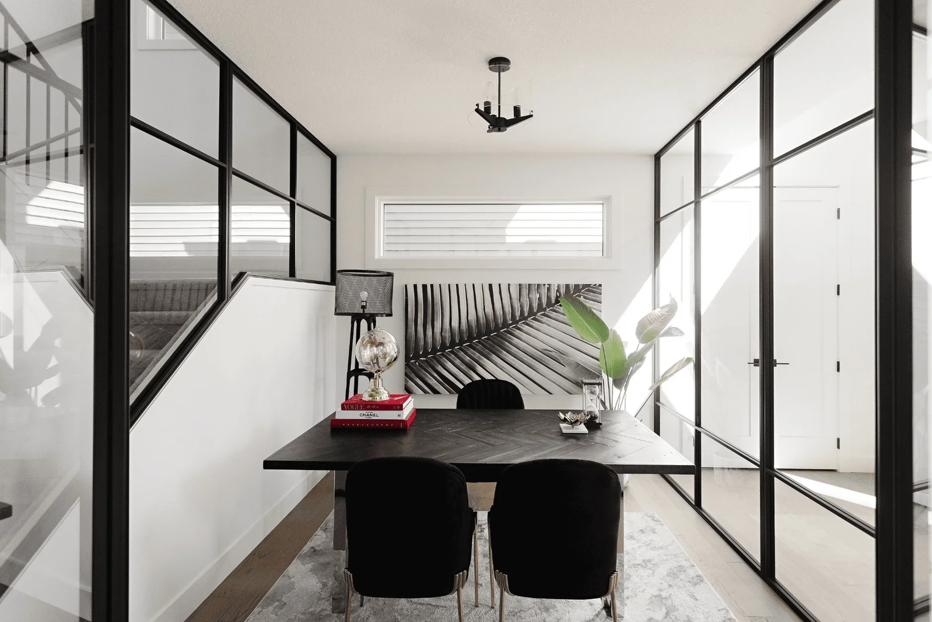 picture of a home office enclosed by black framed glass walls featuring a dark wood herringbone desk and two black velvet chairs with gold legs