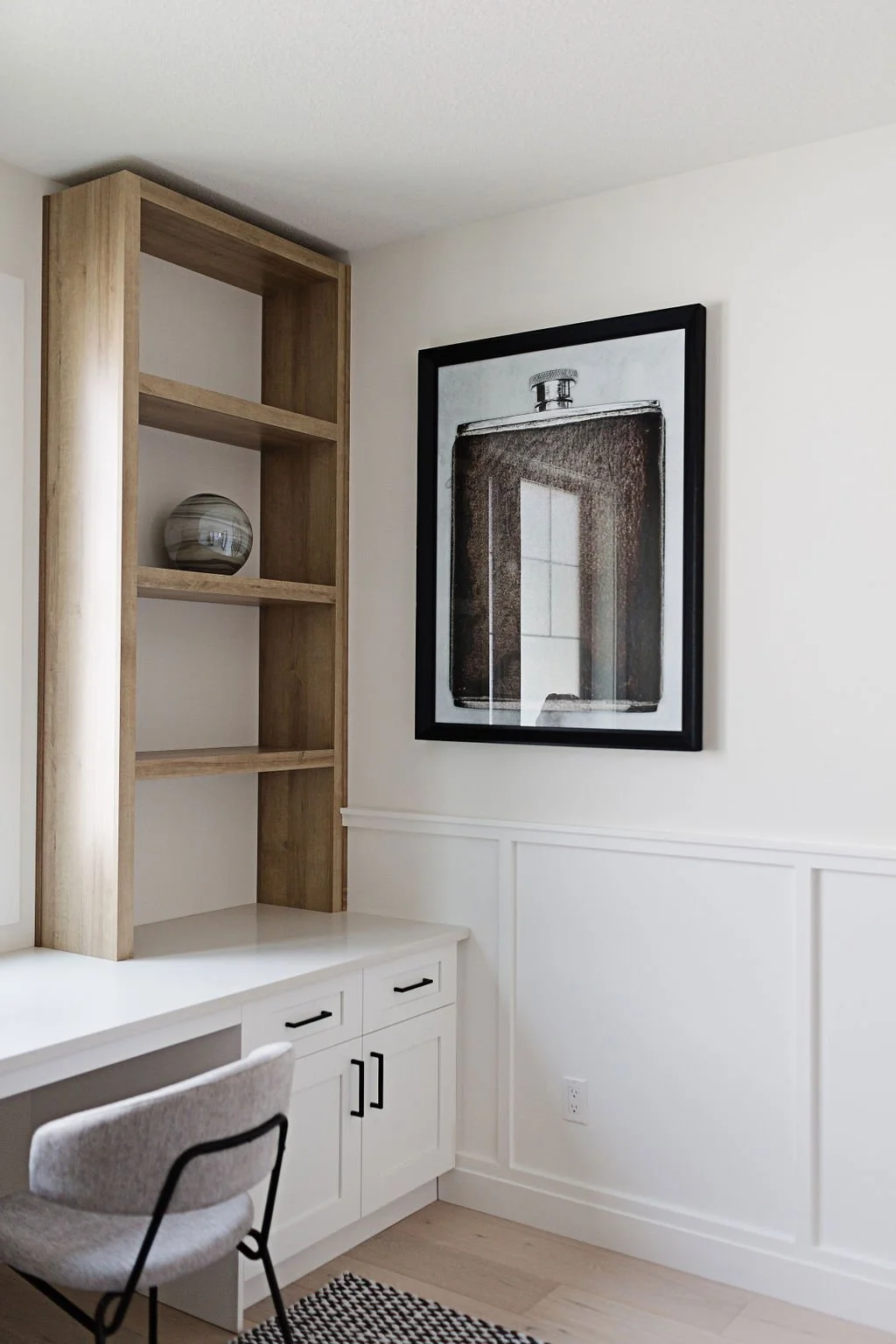 Photo of a grey chair at a white desk with a book shelf above, next to a framed piece of art hanged on a wall with wall moulding and wall trim