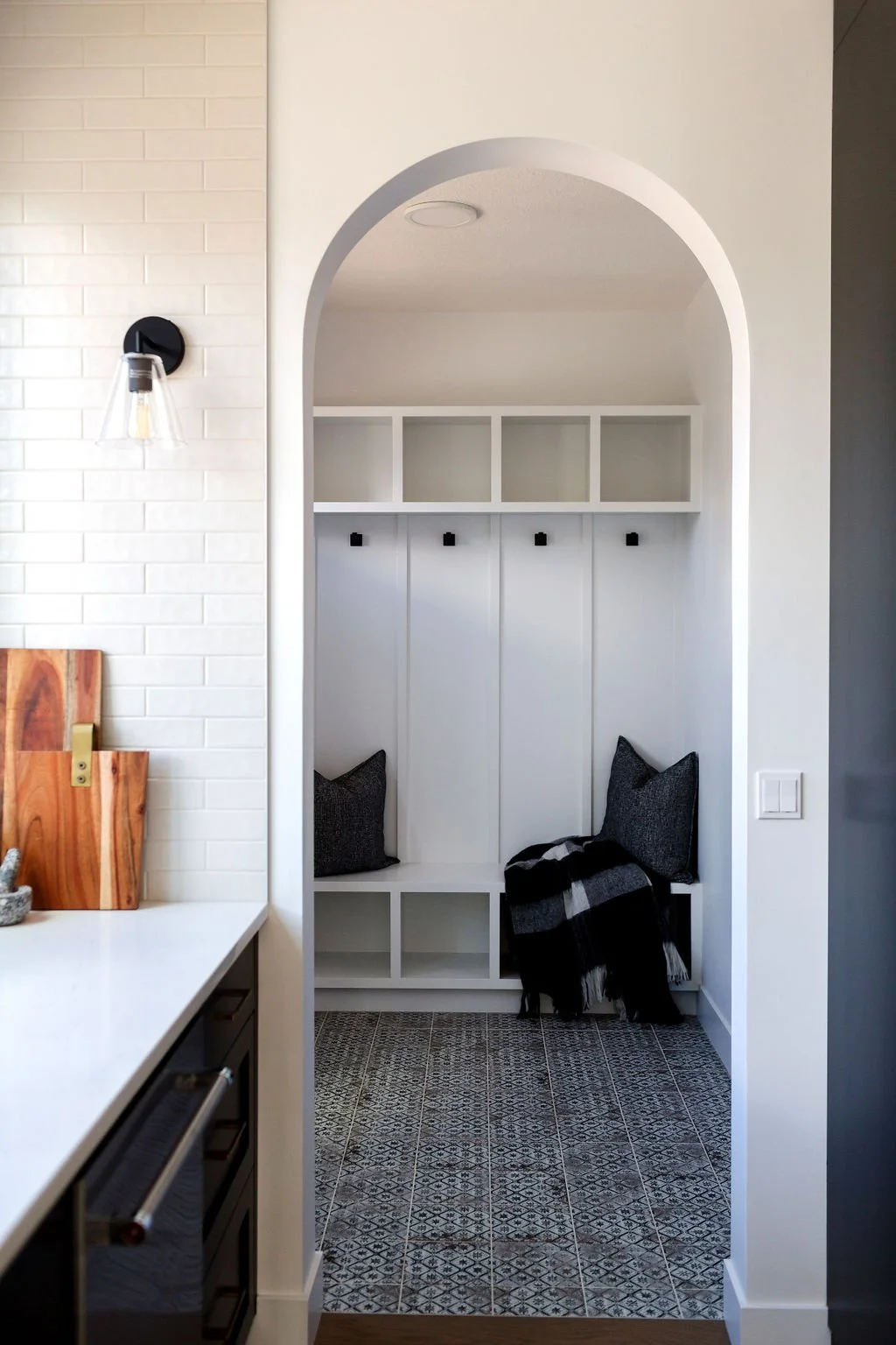 Picture of an arched entryway, leading into a white built-in mudroom bench with cubbies and black hooks, set over a patterned grey tile floor