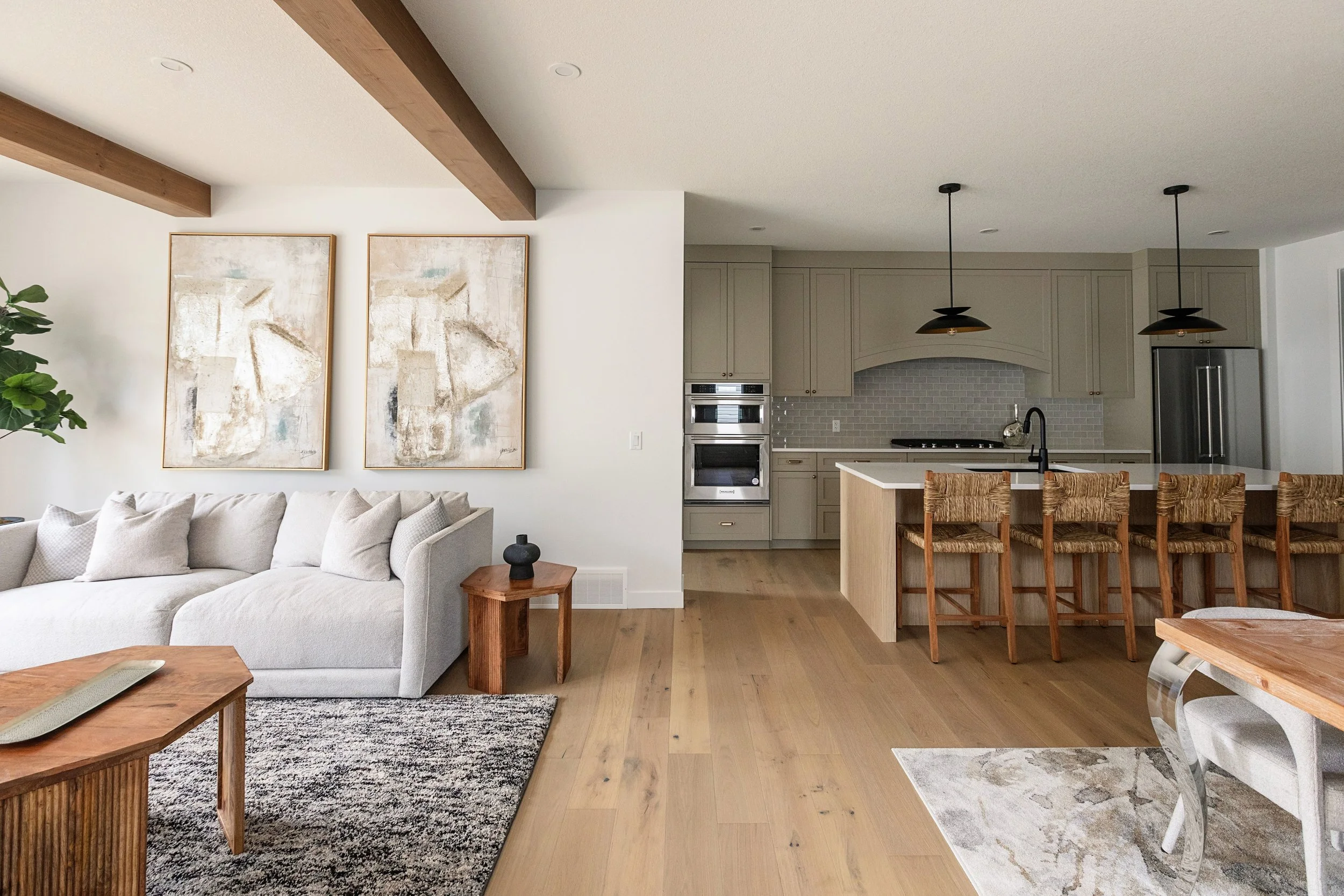 An image of a bright open-concept living room and kitchen with a light sofa, warm wood flooring, large abstract artwork, woven bar stools, and modern pendant lighting