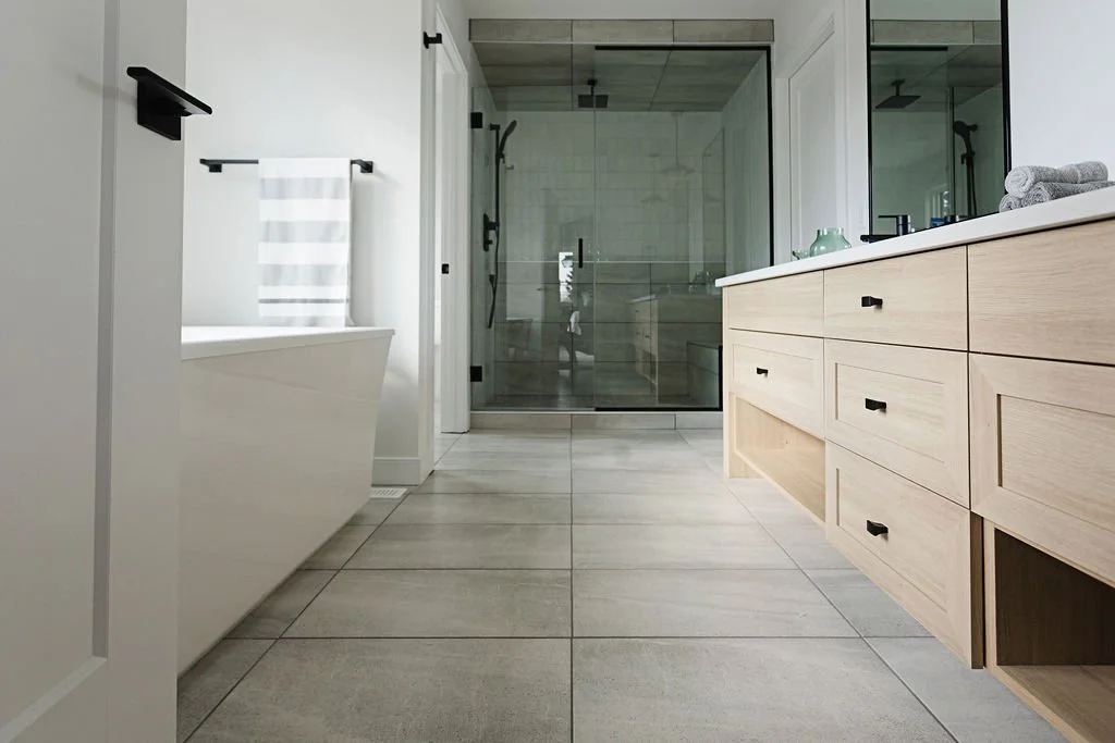 Picture of a modern bathroom showcasing a light wood double vanity with black hardware and large grey floor tiles leading toward a glass enclosed walk-in shower