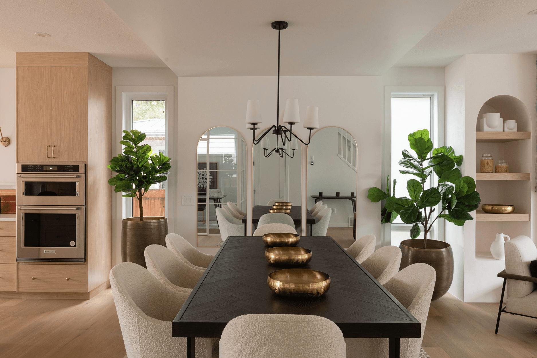 Photo of a black dining table and white chairs in front of a white wall with mirrors next to houseplants