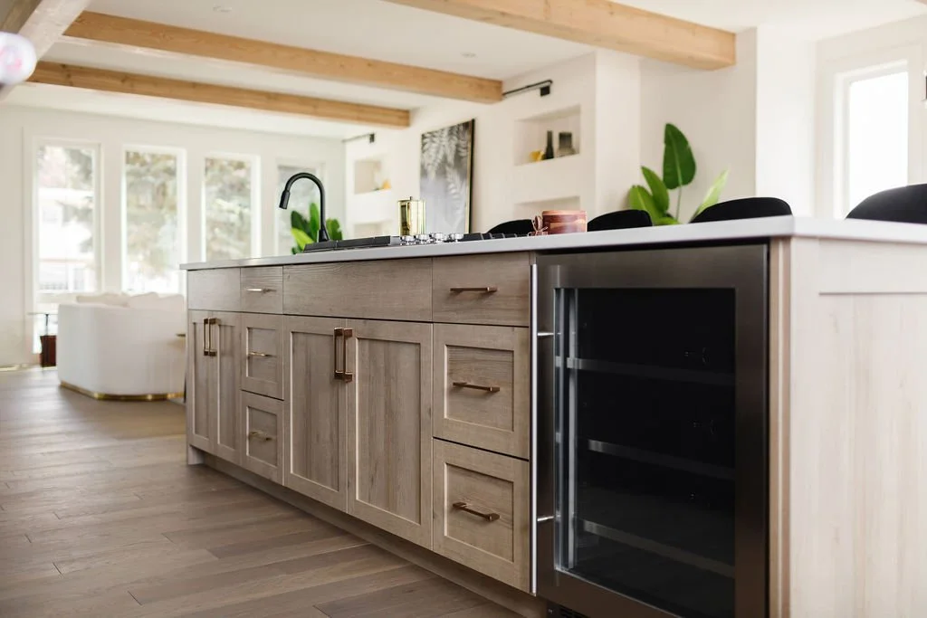 Picture of a kitchen island made of light wood with a built-in wine cooler and integrated drawers, set against a backdrop of white walls and wood ceiling beams