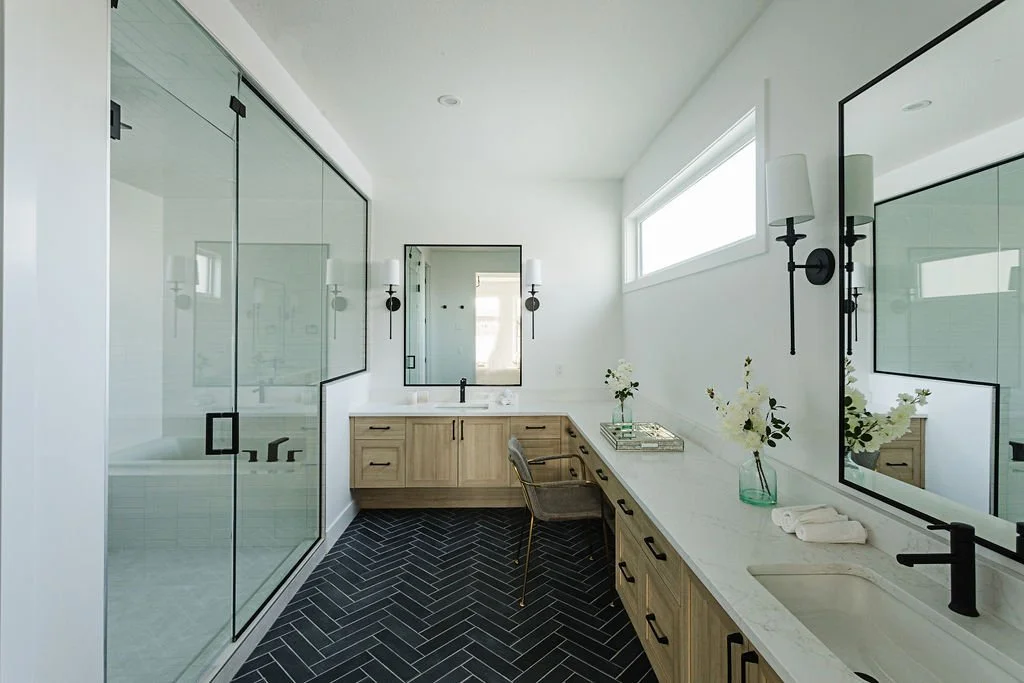 Photo of a large washroom with natural lighting, dark tiled floors, a long L-shaped wood vanity, with several drawers and white countertops, plus two sinks and vanity mirrors facing a glass stand-up shower