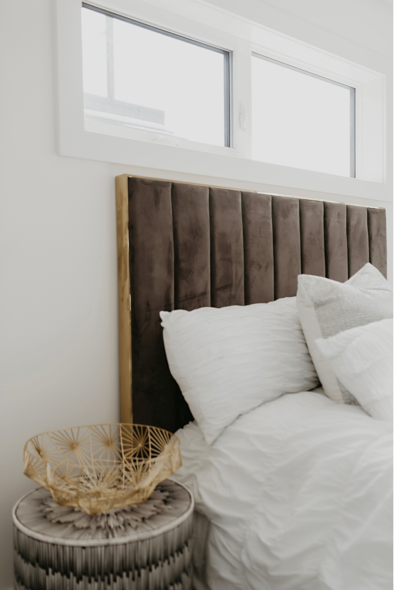 Photo of a grey velvet marshmallow bed frame with white bedding and pillows next to a nightstand with a gold bowl as decor
