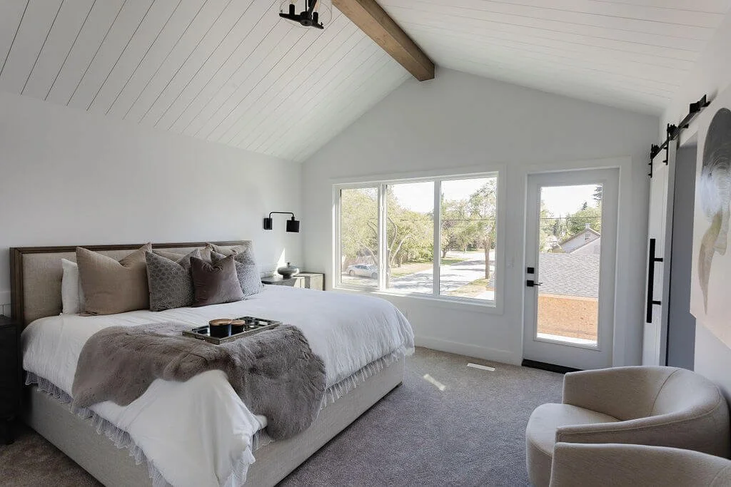Photo of a white bedroom with a white king-sized bed with throw pillows and a blanket next to windows and a glass door