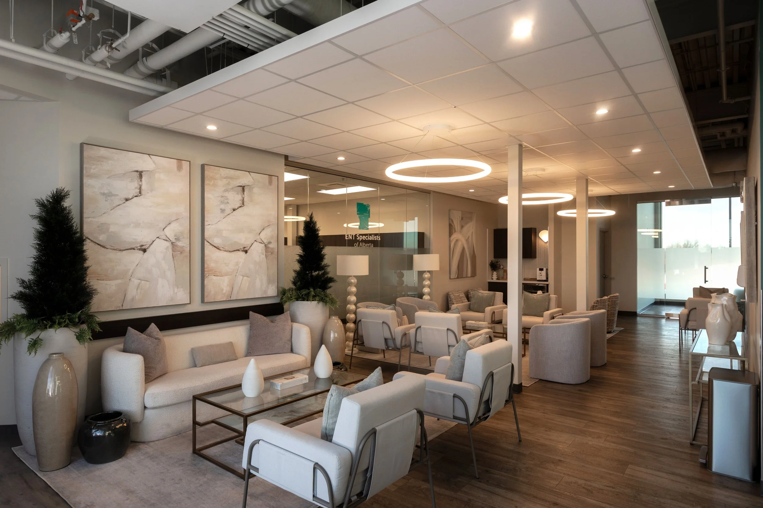 Photo of a waiting room in a clinic with wood floors, matching paintings, white sofas facing armchairs, and bright circular light fixtures above