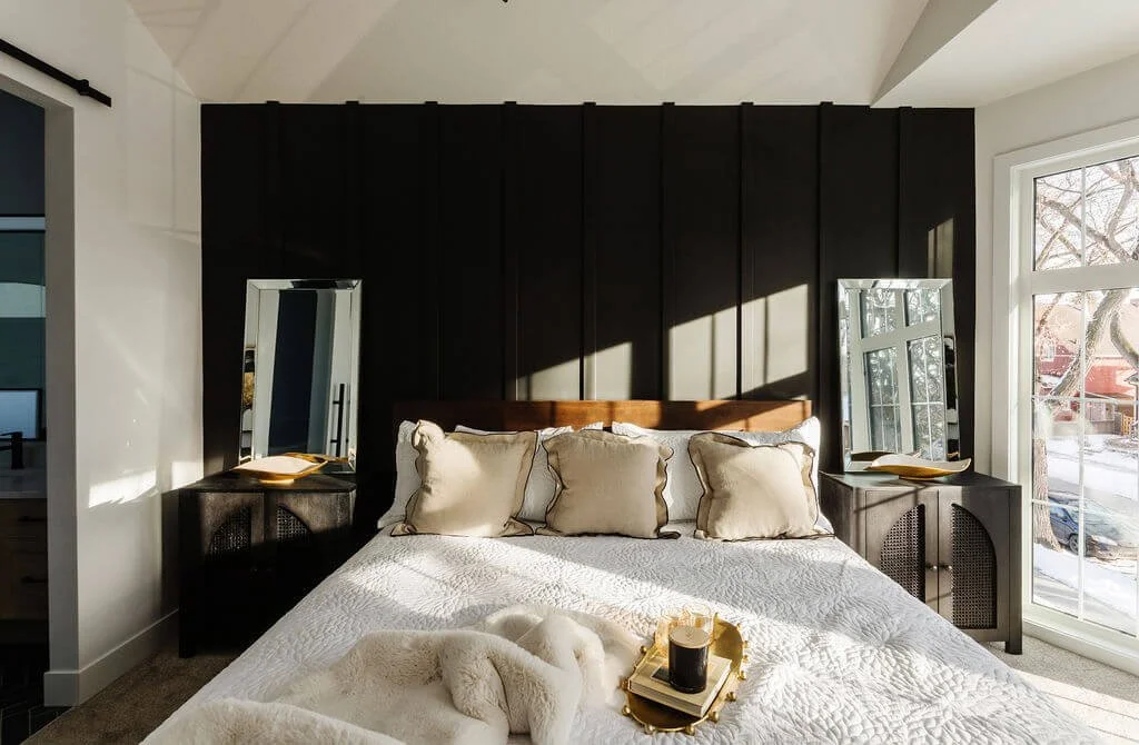 Photo of a black feature wall in a bedroom behind a white queen-sized bed with two mirrors and matching nightstands on either side and blankets a tray and throw pillows on the bed