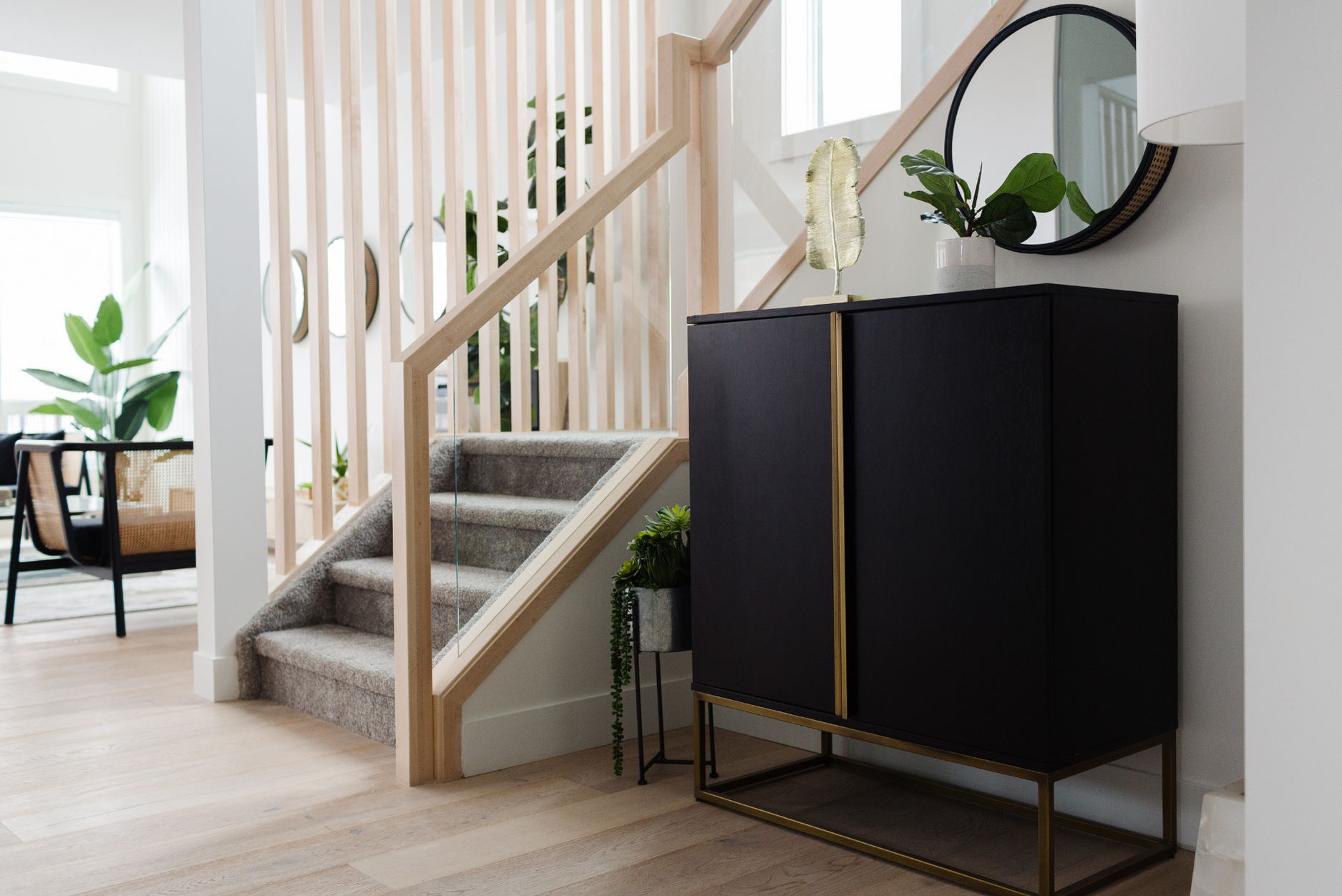 Picture of a bright entryway featuring light wood flooring, a black storage cabinet with gold legs, and a staircase with light wood railings and glass panels