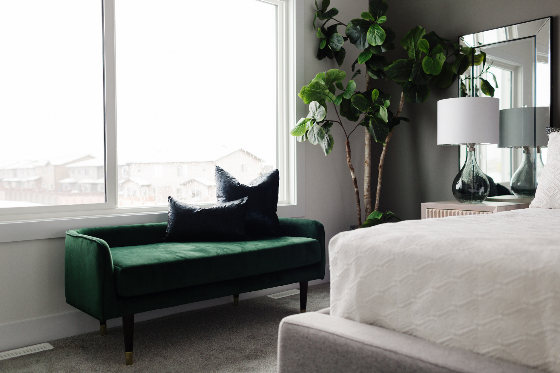 Picture of a cozy corner by a large window, holding a deep green velvet bench and a tall fiddle-leaf fig tree next to a mirrored bedside table
