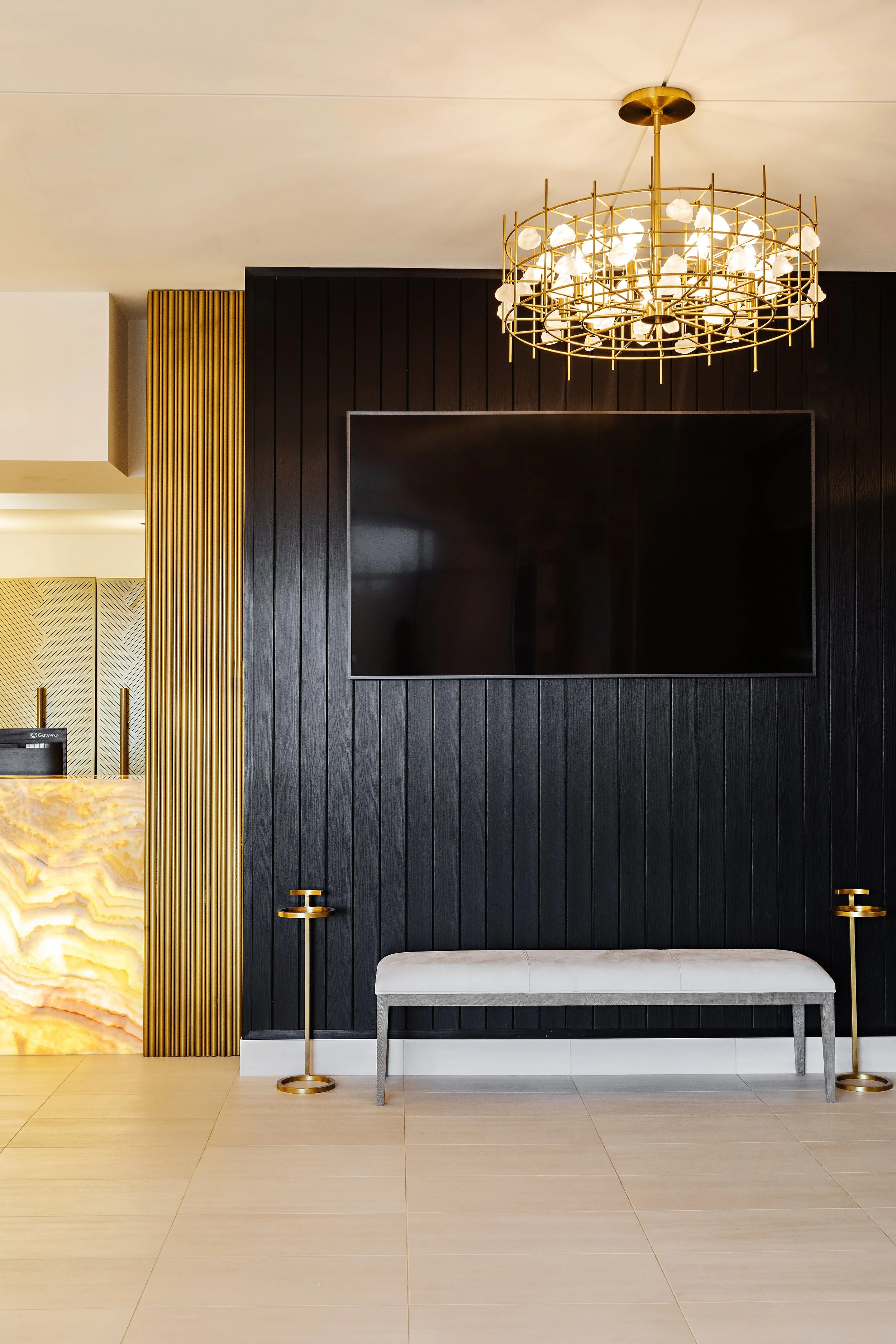 Picture of a gold and black interior design featuring gridded accent walls, a digital screen, a sculptural gold chandelier, twin gold display stands, and a white accent chair