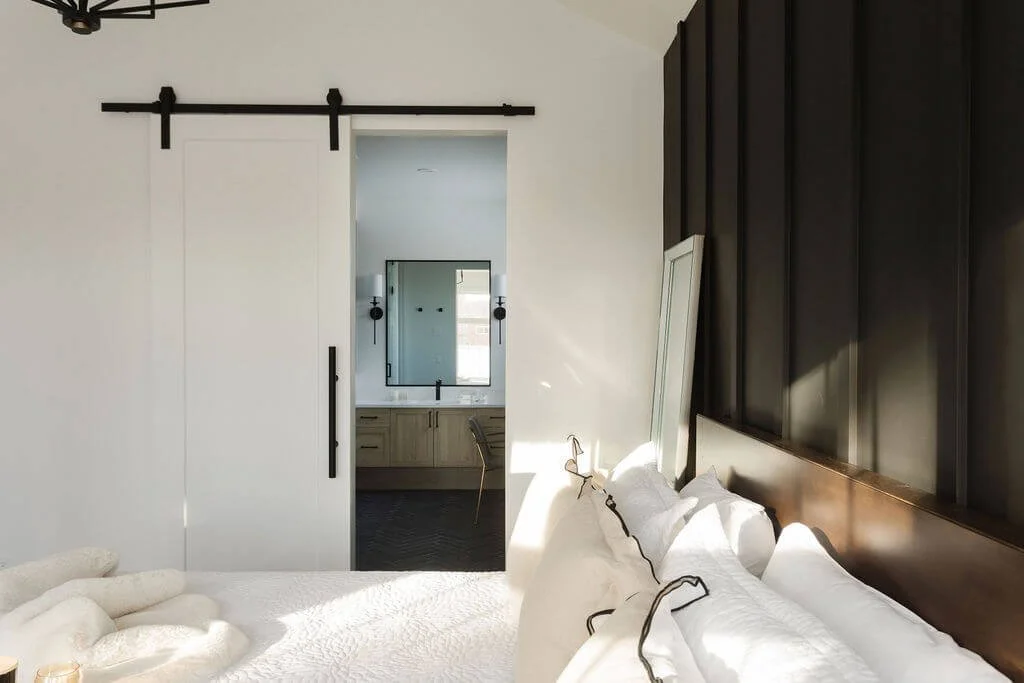 A photo of a modern bedroom with white bedding, a dark accent wall, and a sliding barn interior door styles leading to a bright ensuite