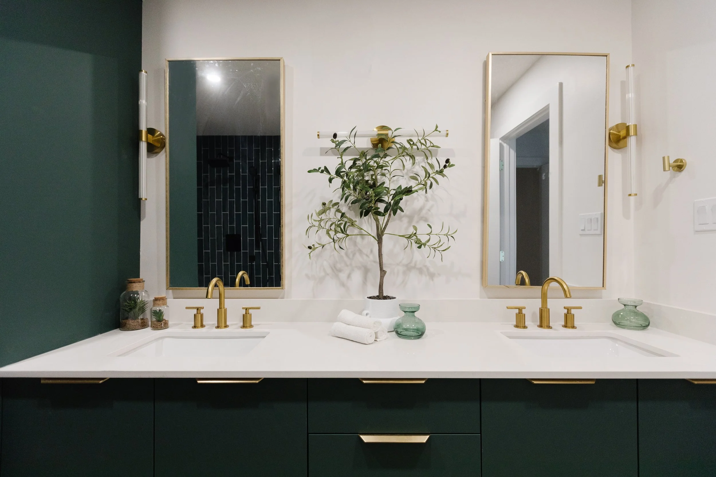 Photo of dark bathroom ideas with a forest green-coloured vanity, gold fixtures, gold rimmed vanity mirrors, and white countertops decorated with houseplants