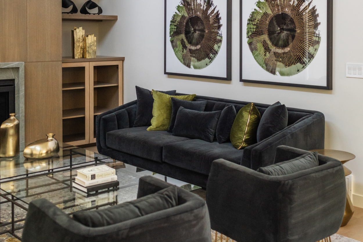 Photo of a black sofa with green throw pillows beneath two identical artworks and next to a glass coffee table and wood shelves