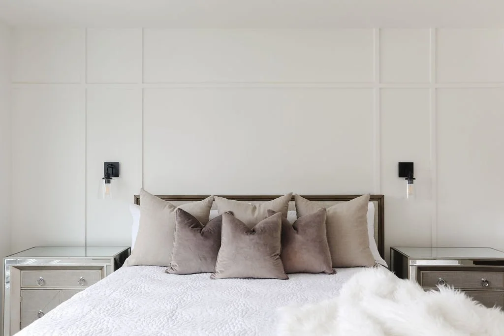 Photo of white wall moulding in a bedroom with two black wall lamps above matching nightstands and a queen-sized white bed with brown throw pillows