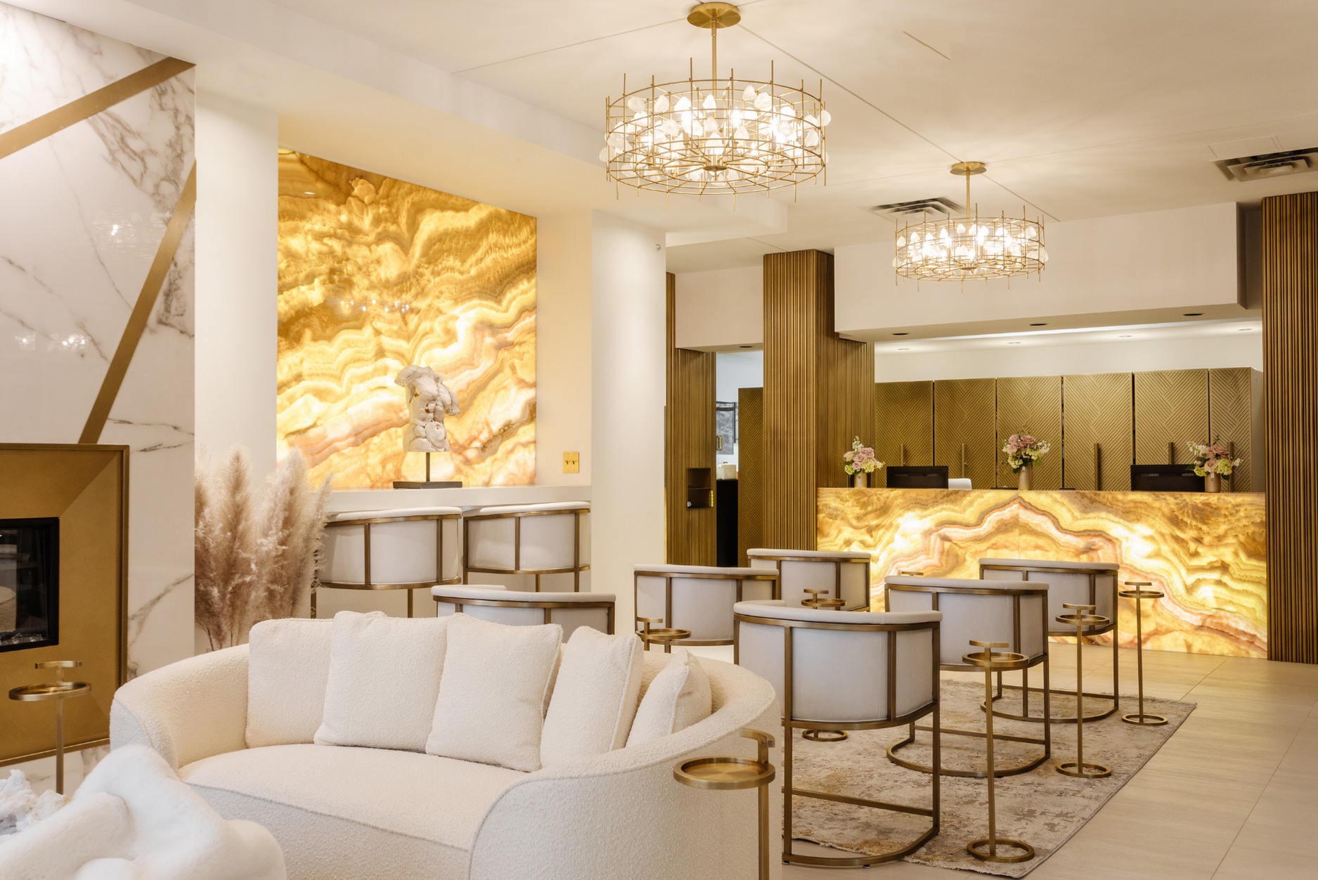 A photo of a luxury interior waiting room featuring white and gold chairs facing gold marble stone walls with bright lighting.