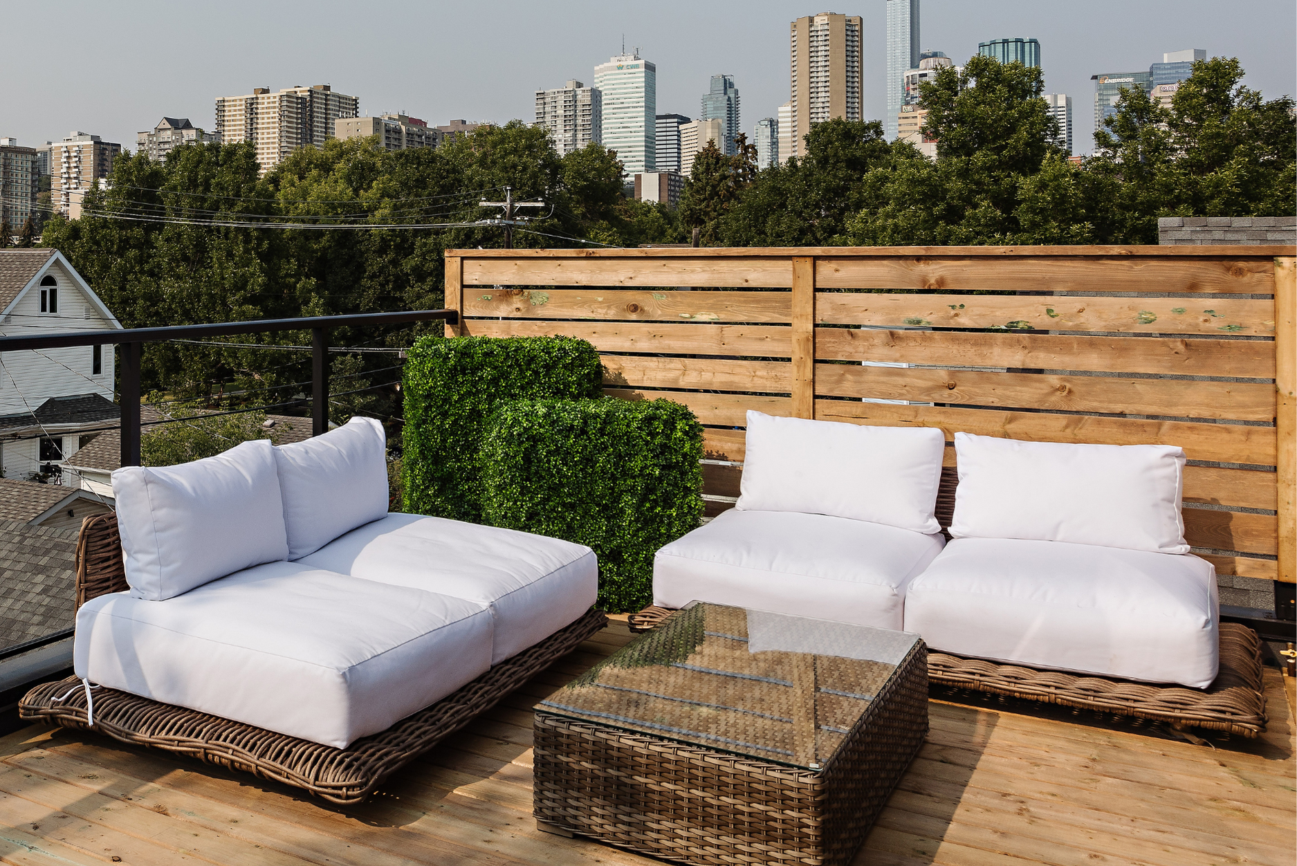 Photo of a wood rooftop deck with white outdoor sofa and fake green bushes with a view of the city behind