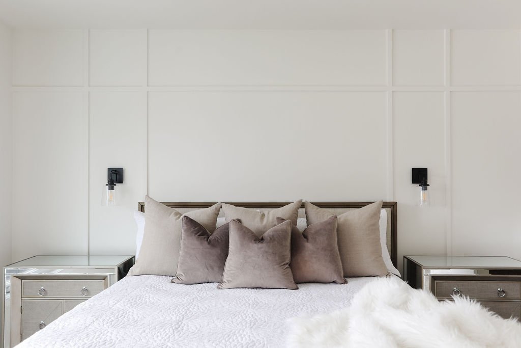 Photo of white wall moulding in a bedroom with two black wall lamps above matching nightstands and a queen-sized white bed with brown throw pillows
