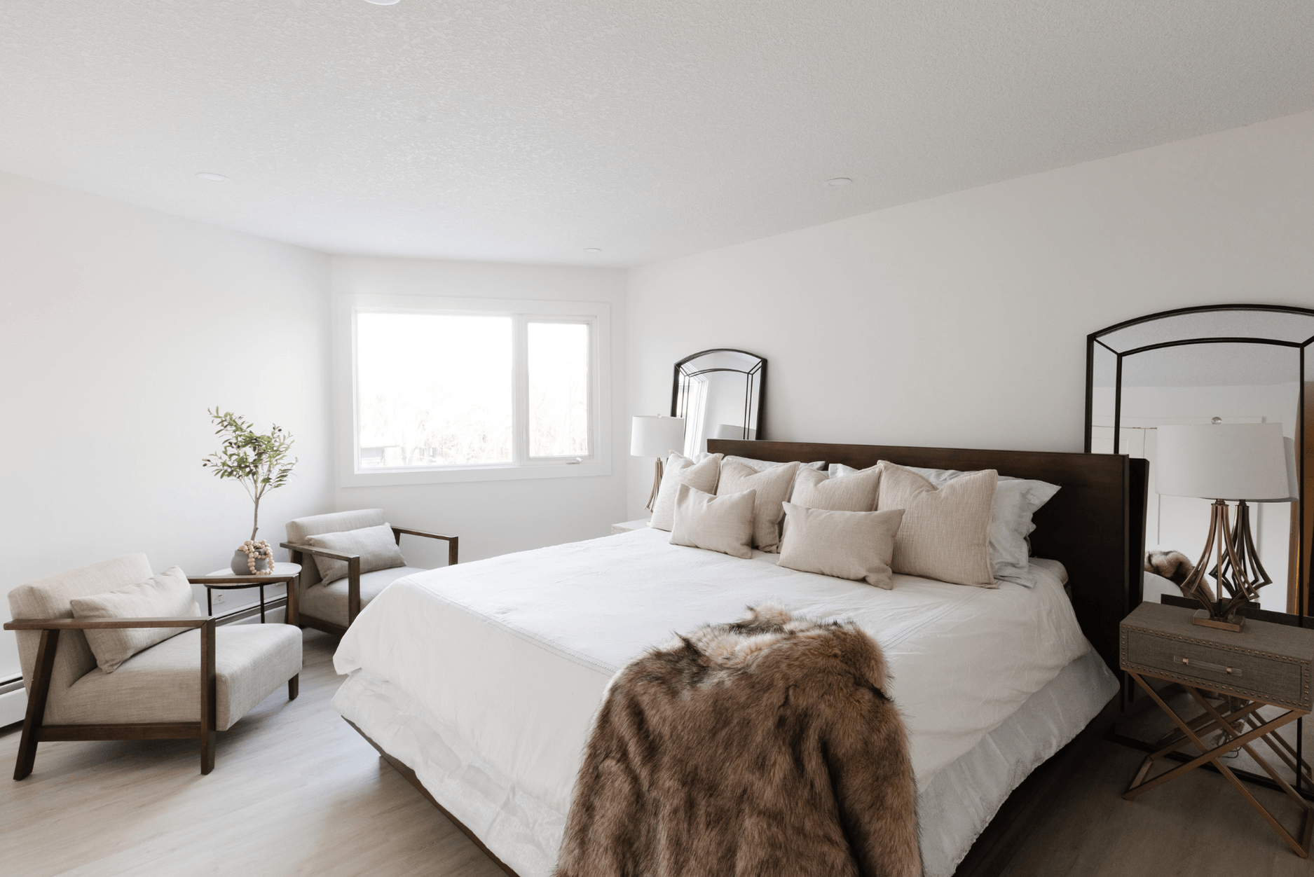 A photo of a bright modern bedroom with a dark wood bed frame, neutral pillows, two beige armchairs by the window, and a faux fur throw blanket at the foot of the bed