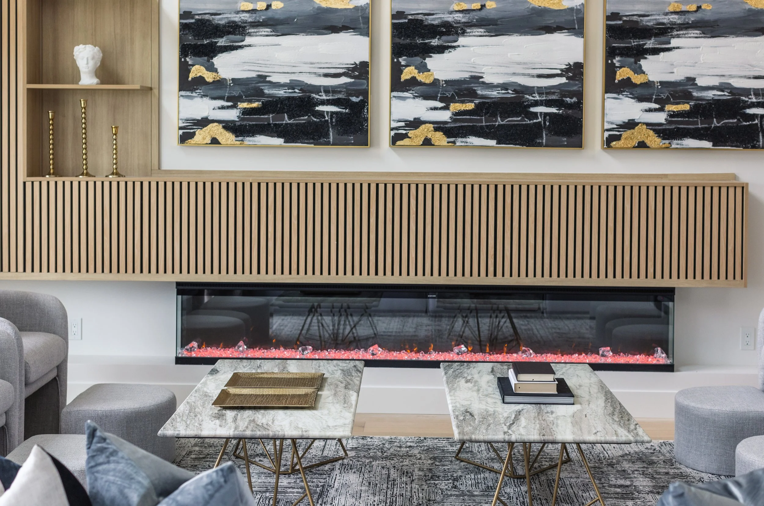 Photo of two identical white marble cofee tables with books and decor surrounded by grey sofas in front of an electric fireplace
