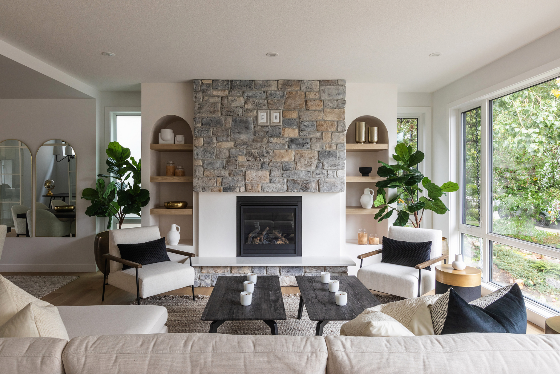 A photo of a bright living room with a stone fireplace, built-in shelves, neutral armchairs, a beige sofa, and black accent pillows