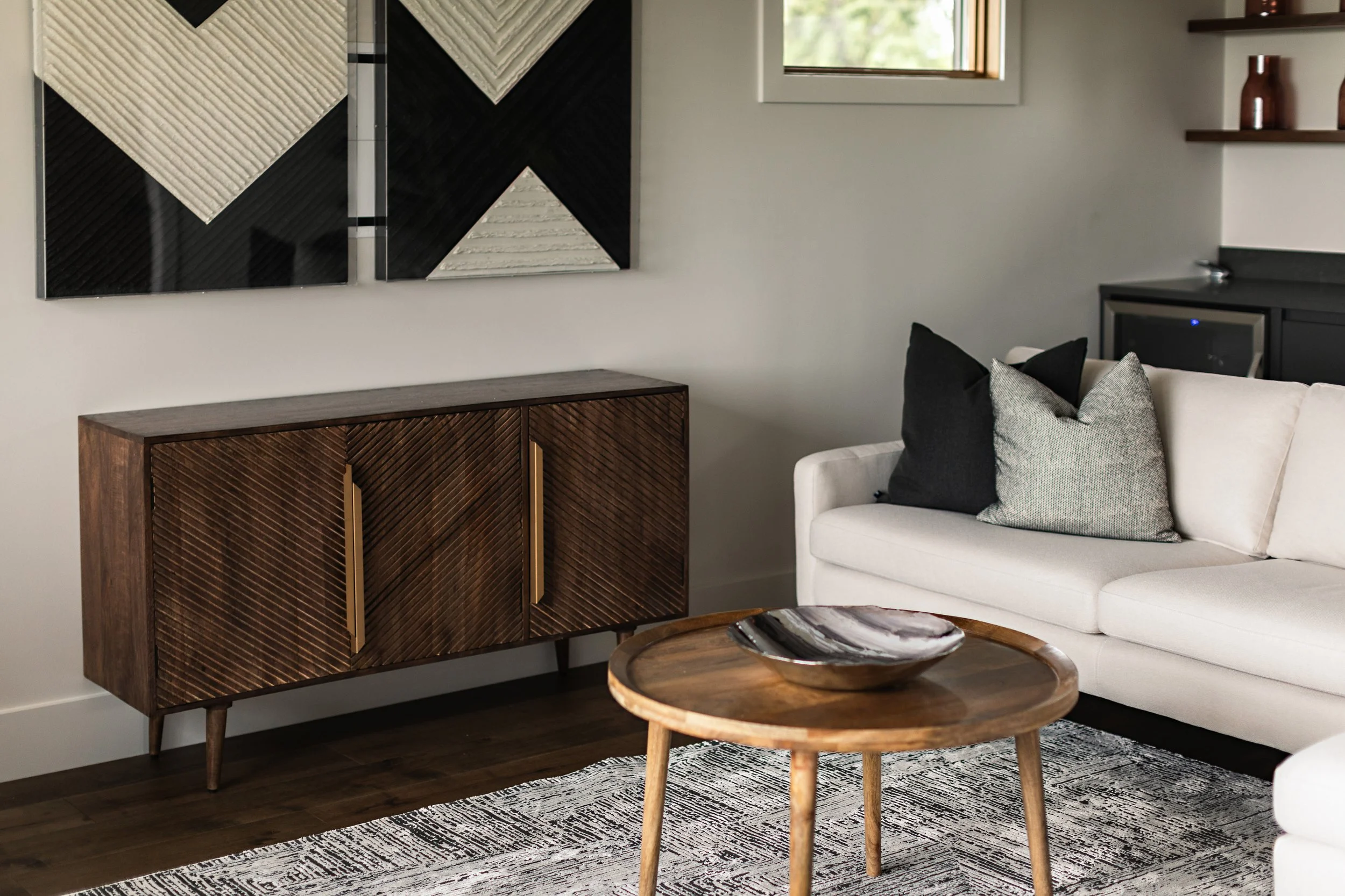 A photo of a cozy living room corner featuring a light sofa, a wooden sideboard, a round wooden coffee table, and black and white abstract wall art