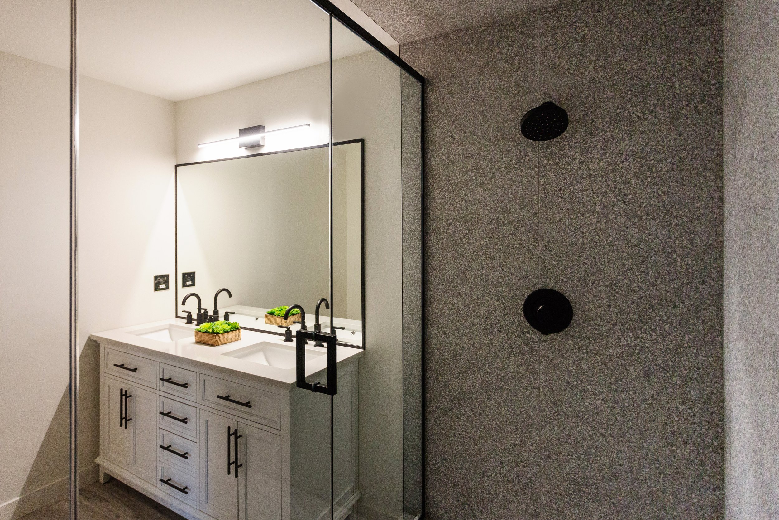 Photo of a bathroom shower with stone walls next to a large square mirror and white countertop with dual sinks